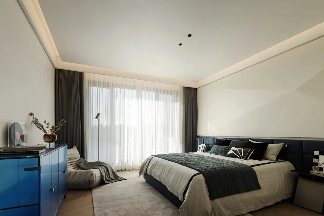 A modern bedroom featuring a large window with sheer curtains, a comfortable bed with a blue leather headboard, and a blue ca
