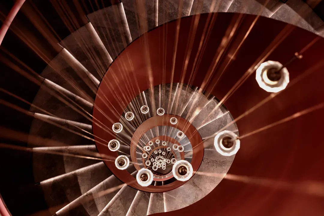 A modern metal staircase spiral upwards, featuring a warm, industrial aesthetic with sleek, angular lines.