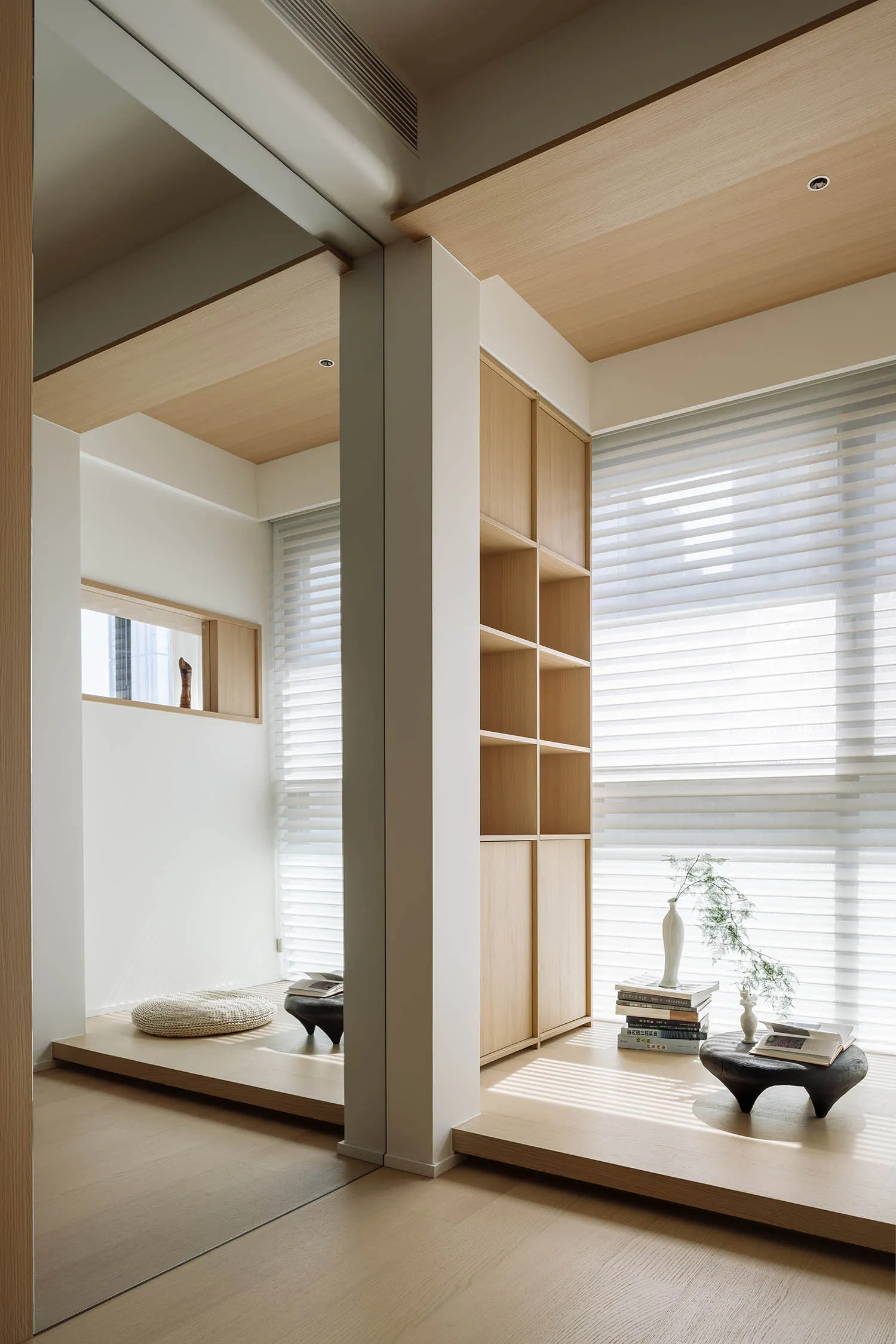 A minimalist living space featuring wooden elements and a sunlit room with a bookshelf and window.