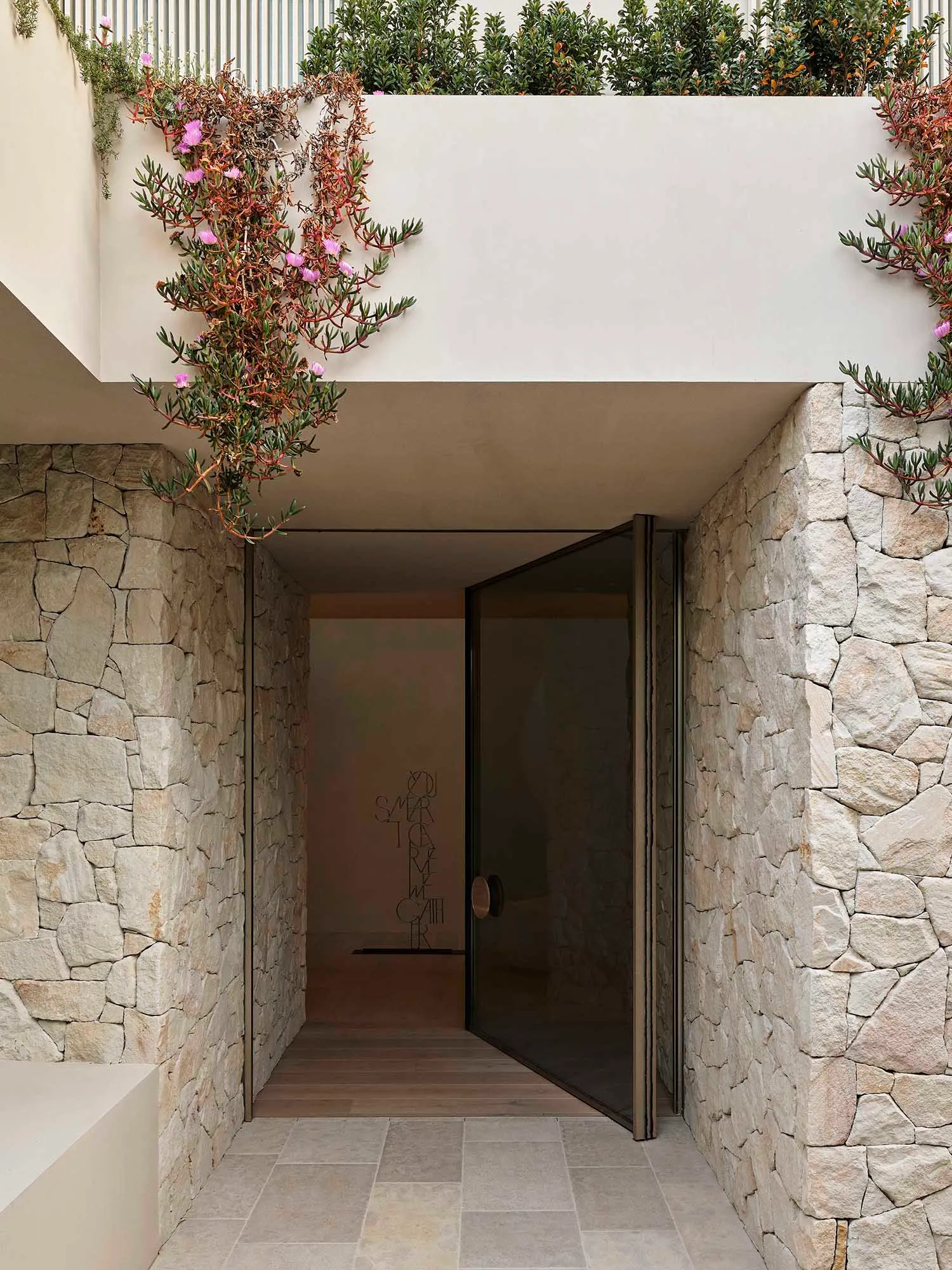 Modern entranceway with stone walls and a glass door, featuring greenery above and a wooden floor.