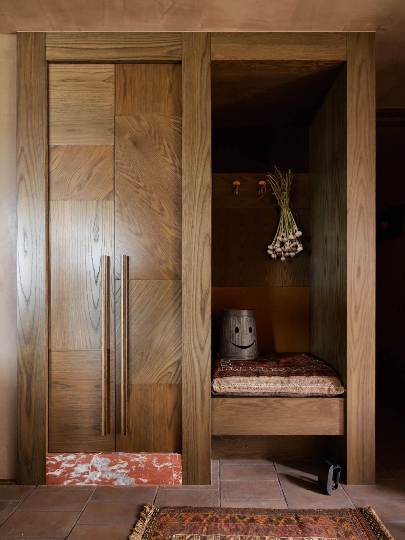 A cloakroom with wooden doors, a bench, and a bunch of onions hanging. Features include a tiled floor and a rustic wooden int