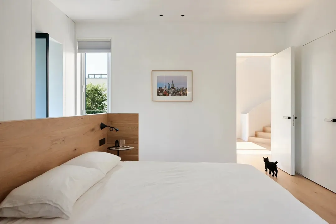 Modern minimalist bedroom with wooden headboard, white bedding, cityscape artwork, and black cat.