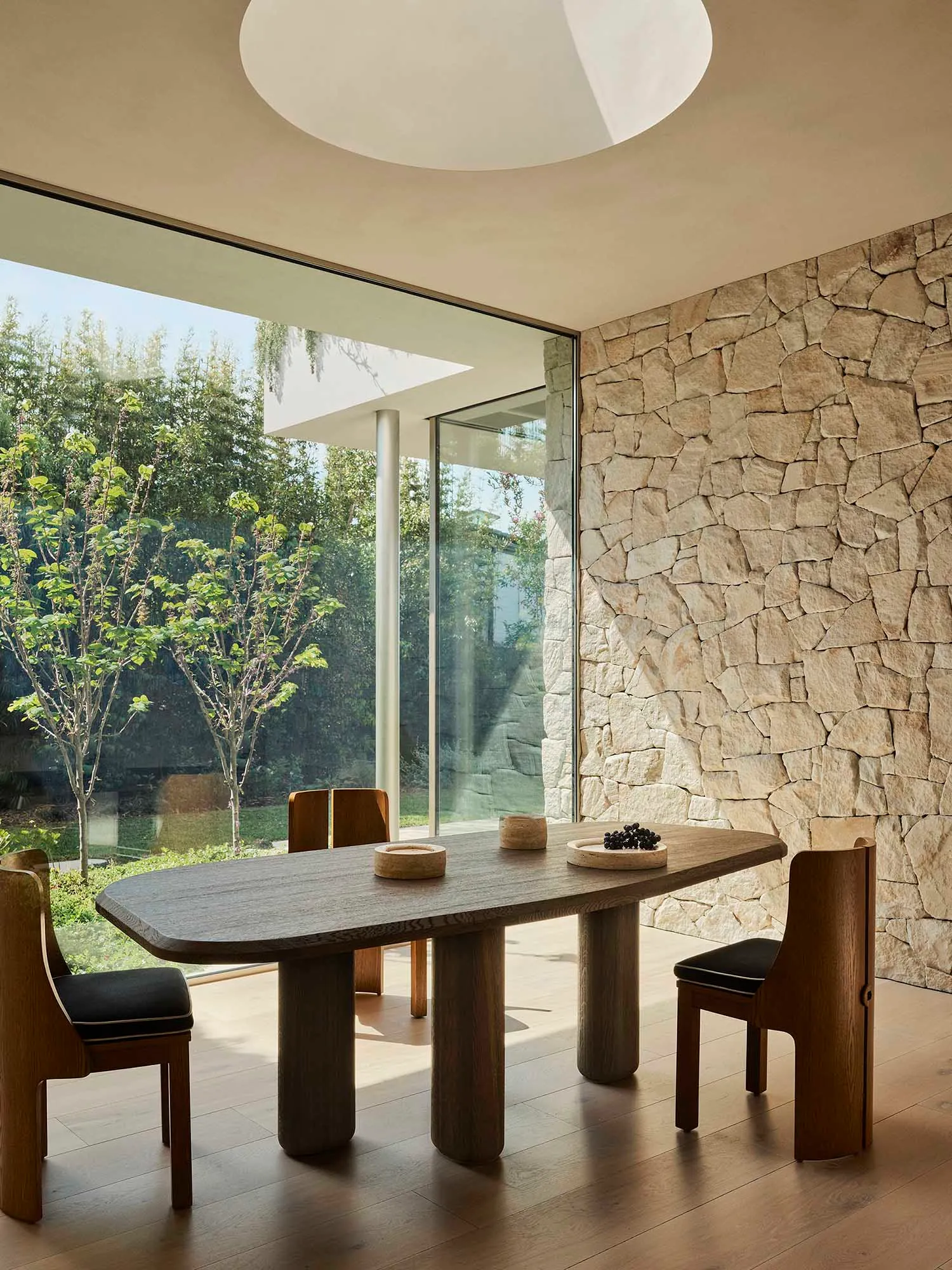 A minimalist dining room with a wooden table and a stone wall, featuring floor-to-ceiling windows that allow natural light to