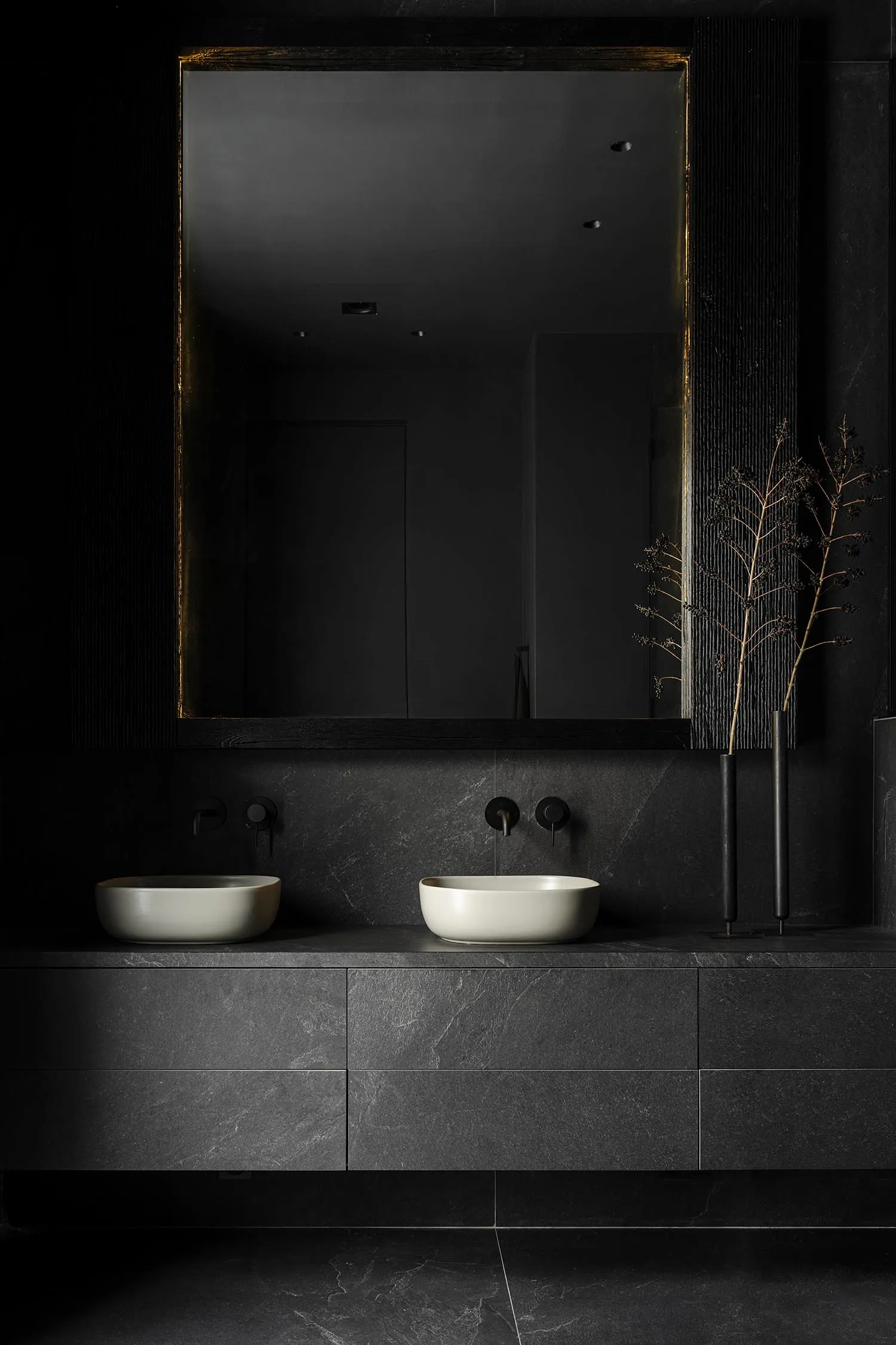 A modern bathroom with dark granite tiles, white vessel sinks, black fixtures, and a black-framed mirror.
