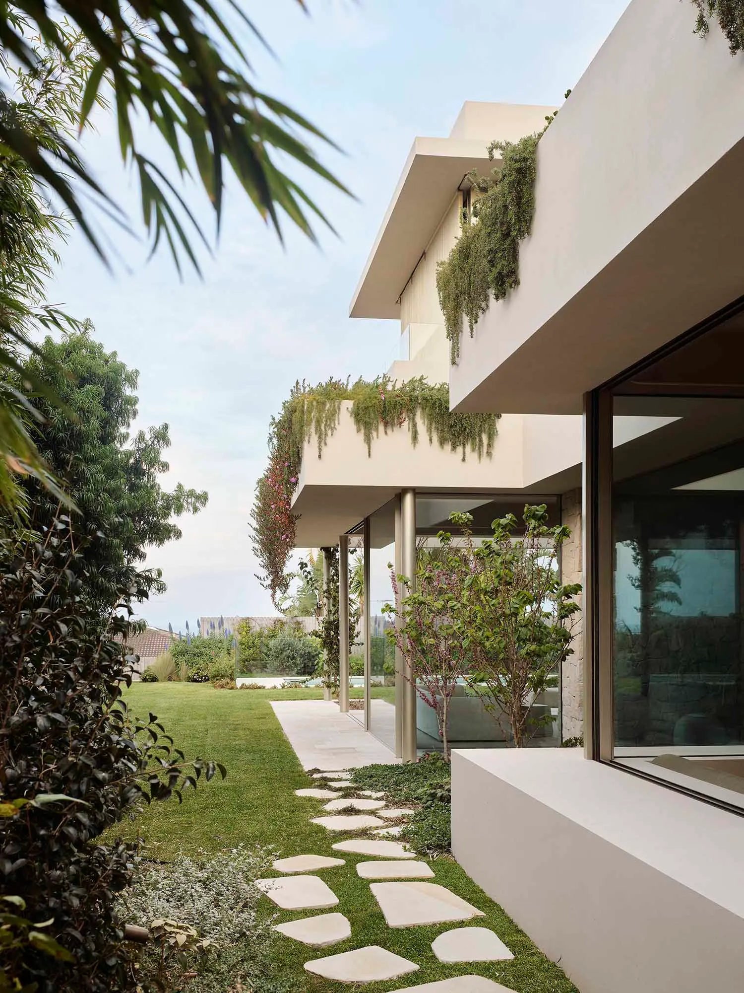A modern residential garden features a path with stepping stones leading to a house with concrete walls, greenery on the balc