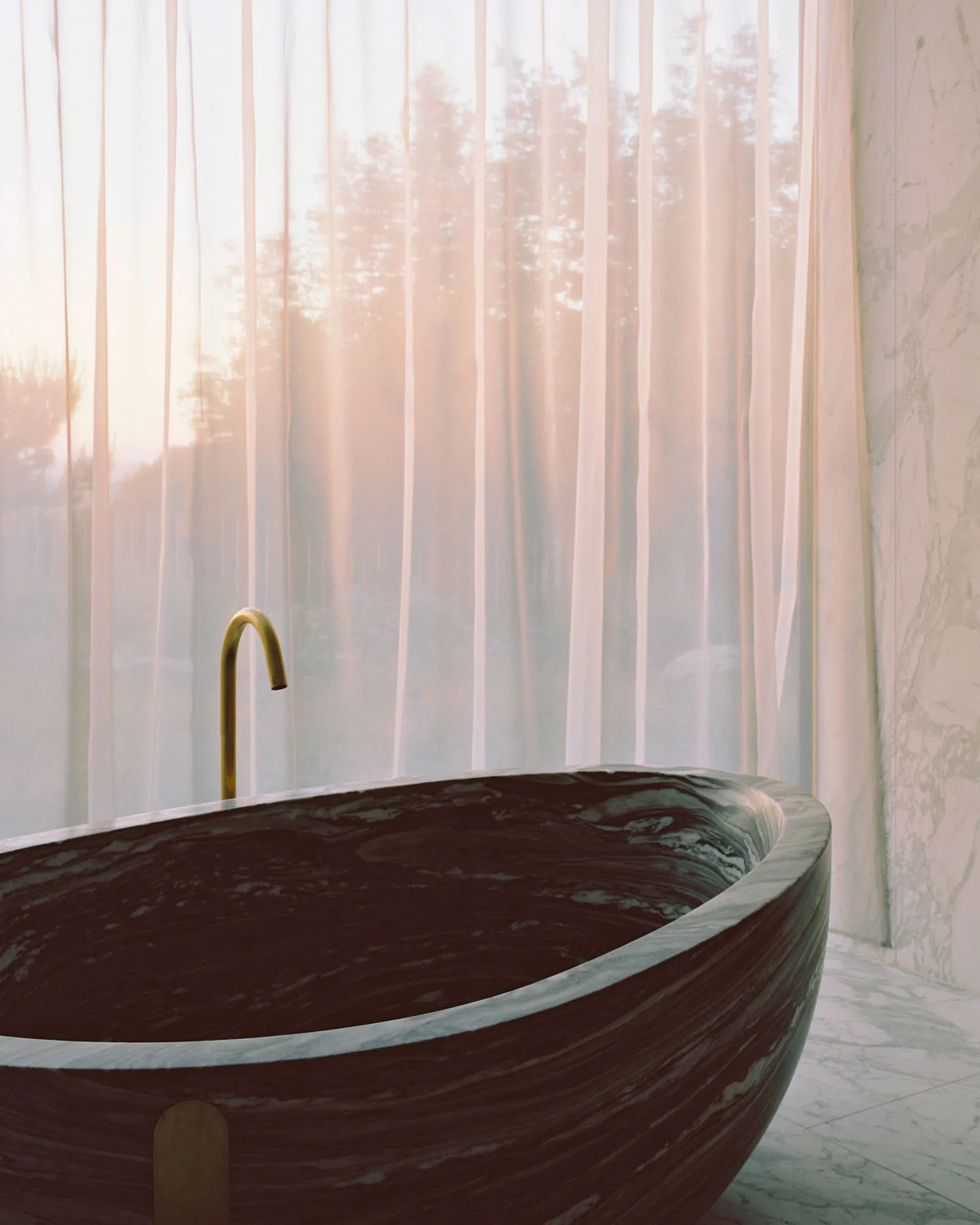 A minimalist bathroom featuring a marble bathtub filled with water, with sheer curtains and a view of a dusk sky outside.