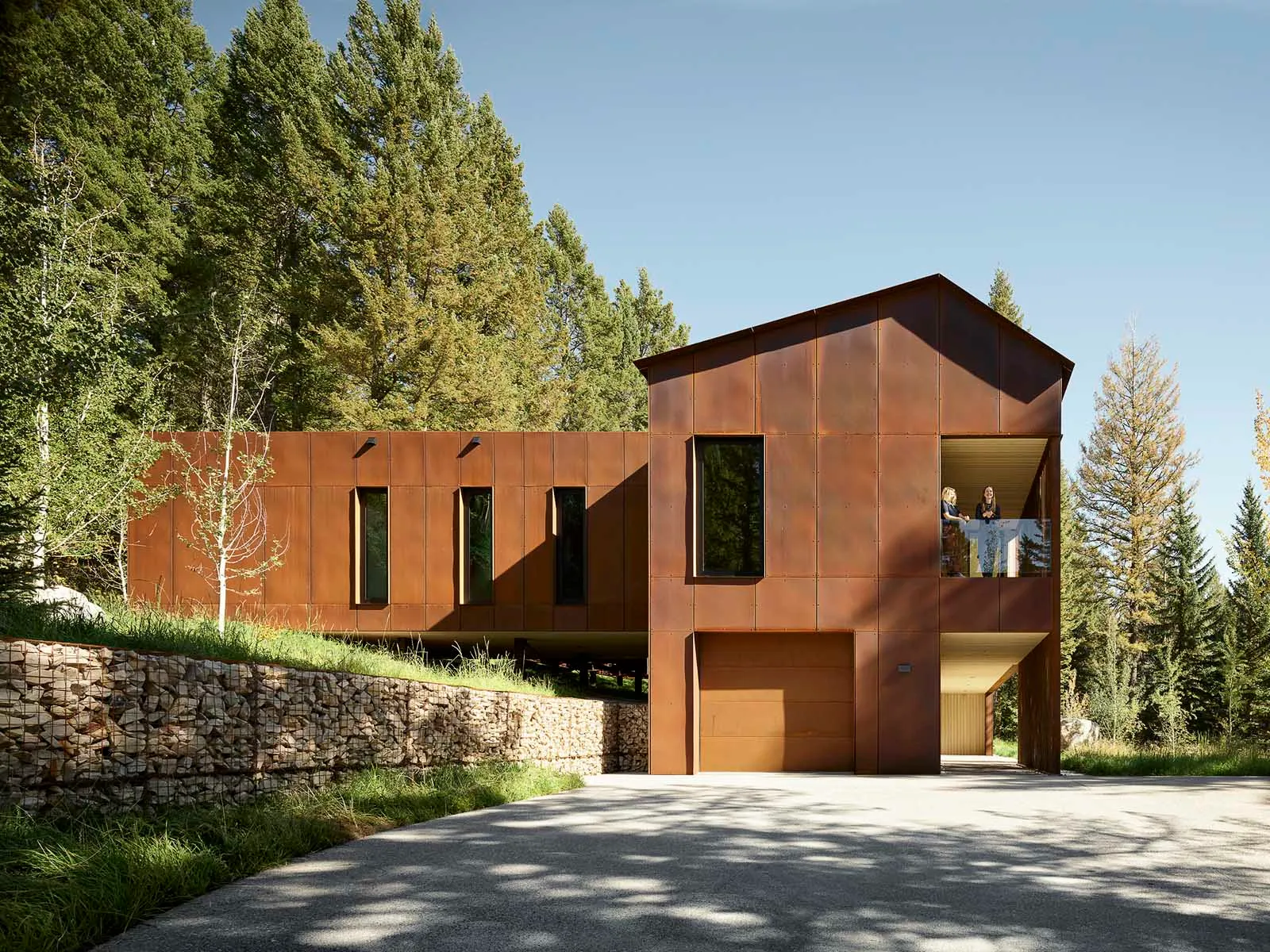 Modern residential house with metal siding, surrounded by trees with a gravel driveway.