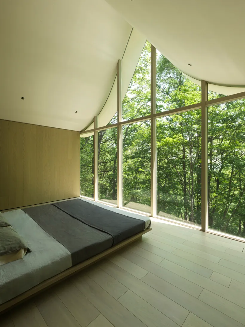 A modern minimalist bedroom with a large window showcasing a forest view, featuring wooden flooring and a low-profile bed.