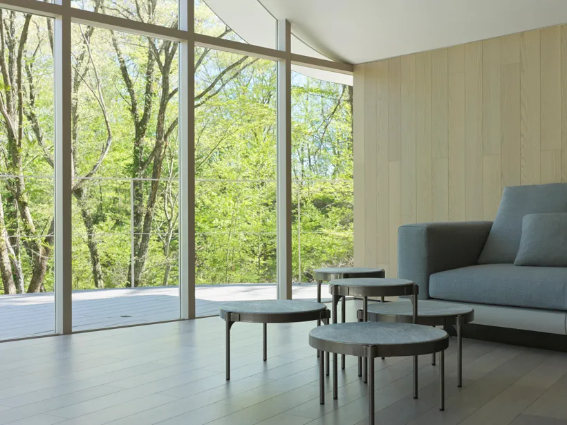 A modern minimalist living room featuring wooden furniture and large windows with a view of greenery.