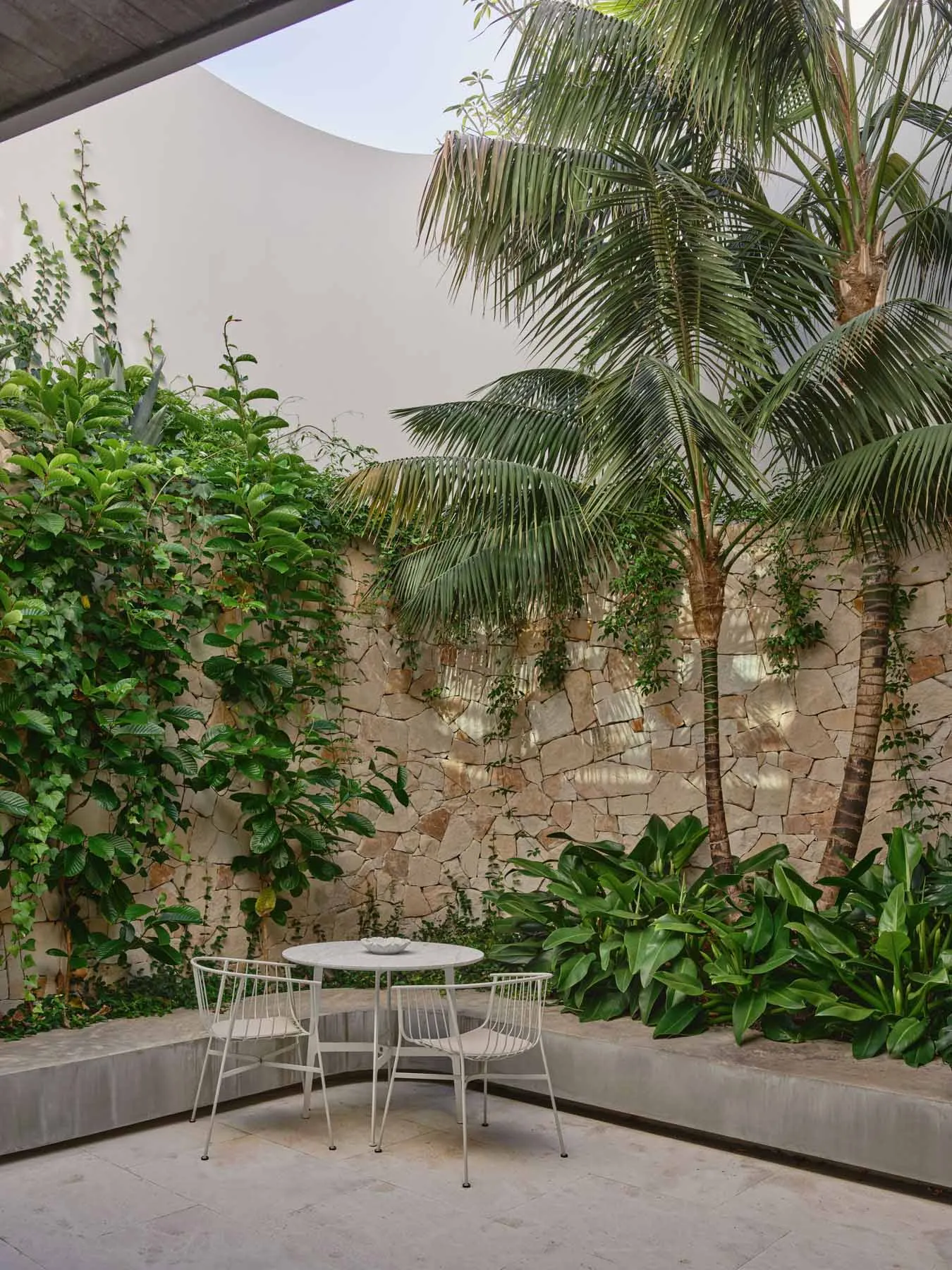 A modern outdoor space featuring a circular white table and chairs on a concrete patio surrounded by greenery and palm trees.