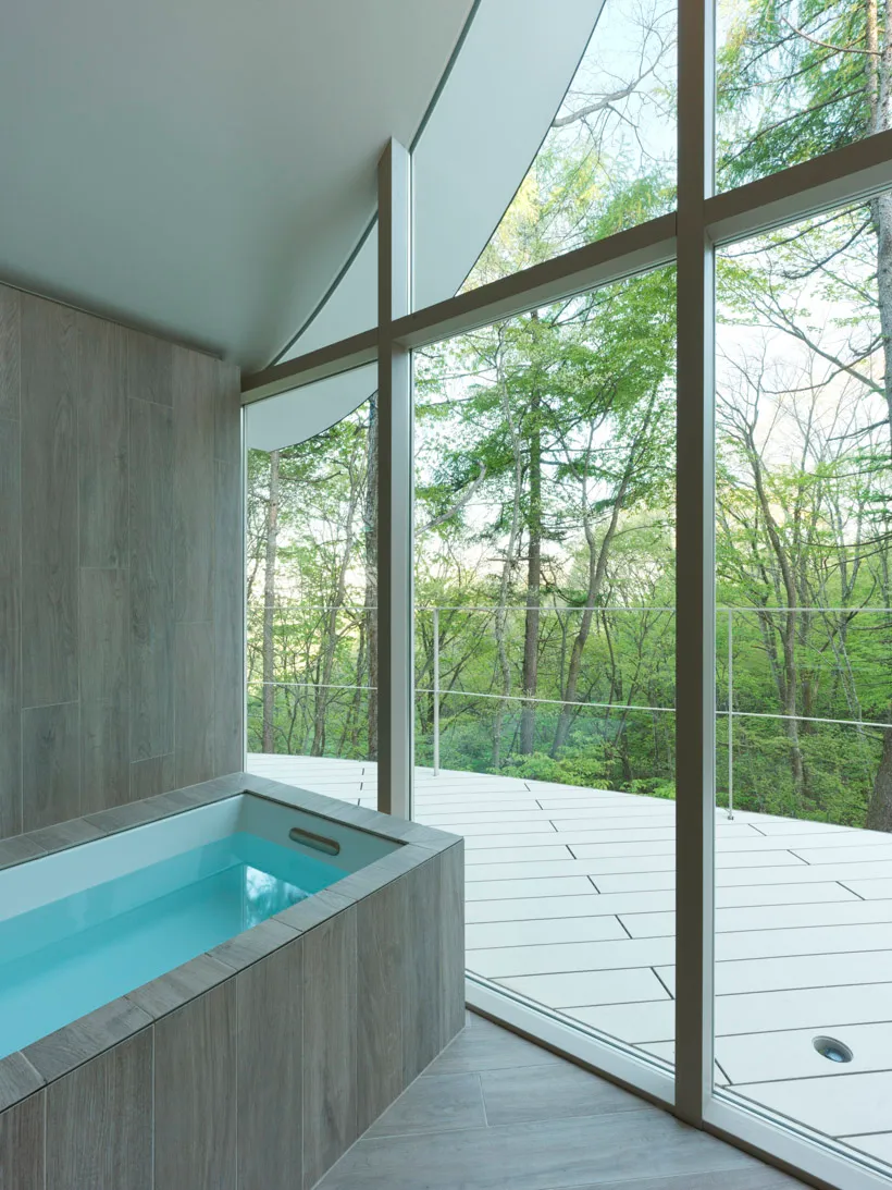 A modern residential bathroom with a glass wall overlooking a forest, featuring minimalist design and a wooden bathtub.
