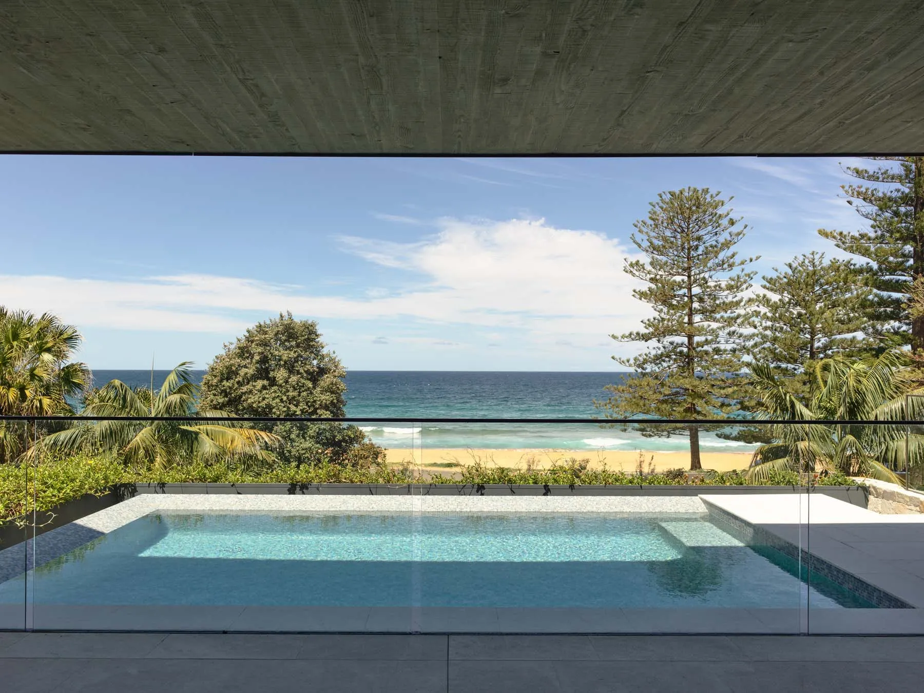 A modern swimming pool overlooking the ocean, surrounded by lush greenery and a concrete patio.