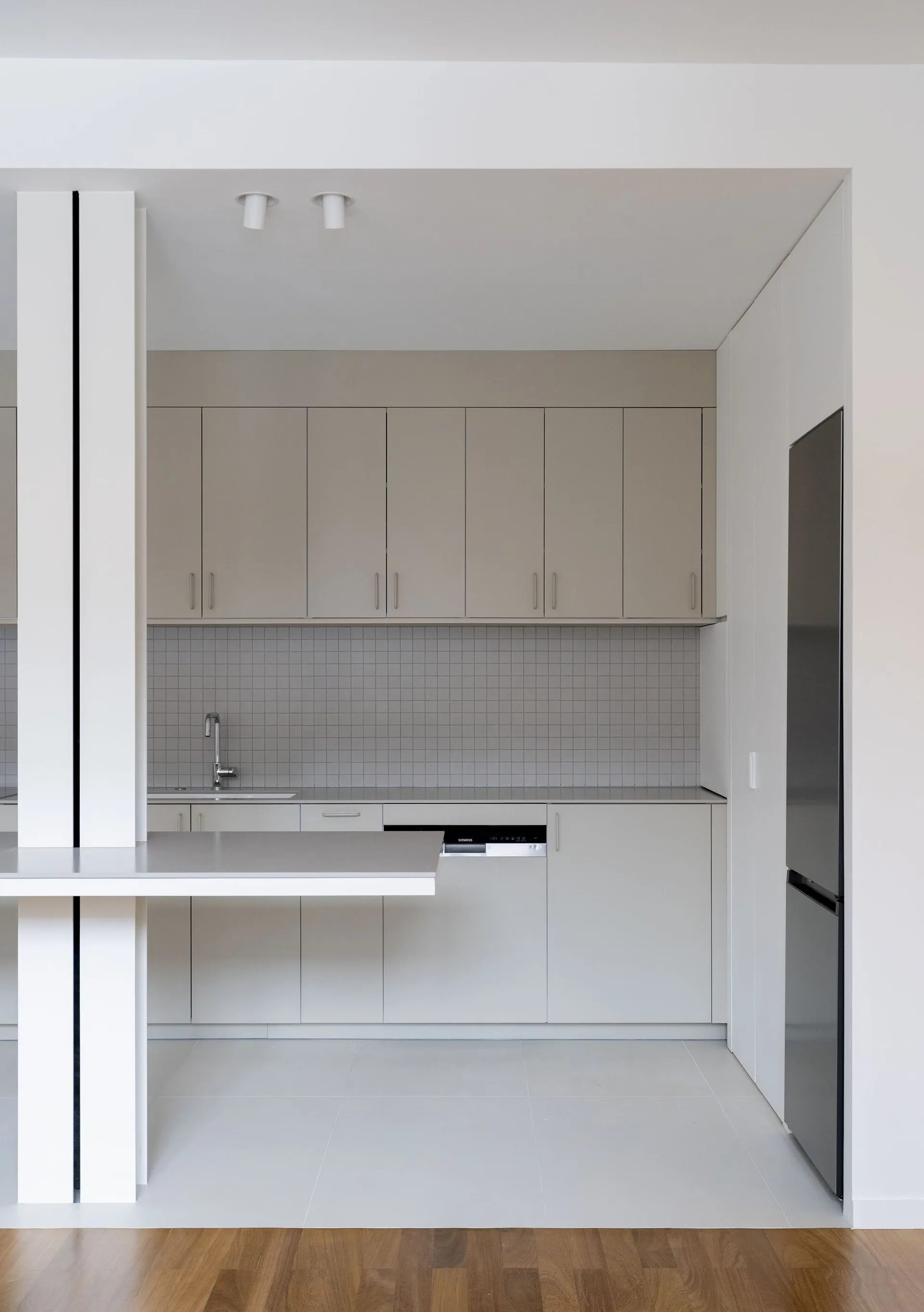 A modern, minimalist residential kitchen with light grey cabinets, a white countertop, and a backsplash made of small, square