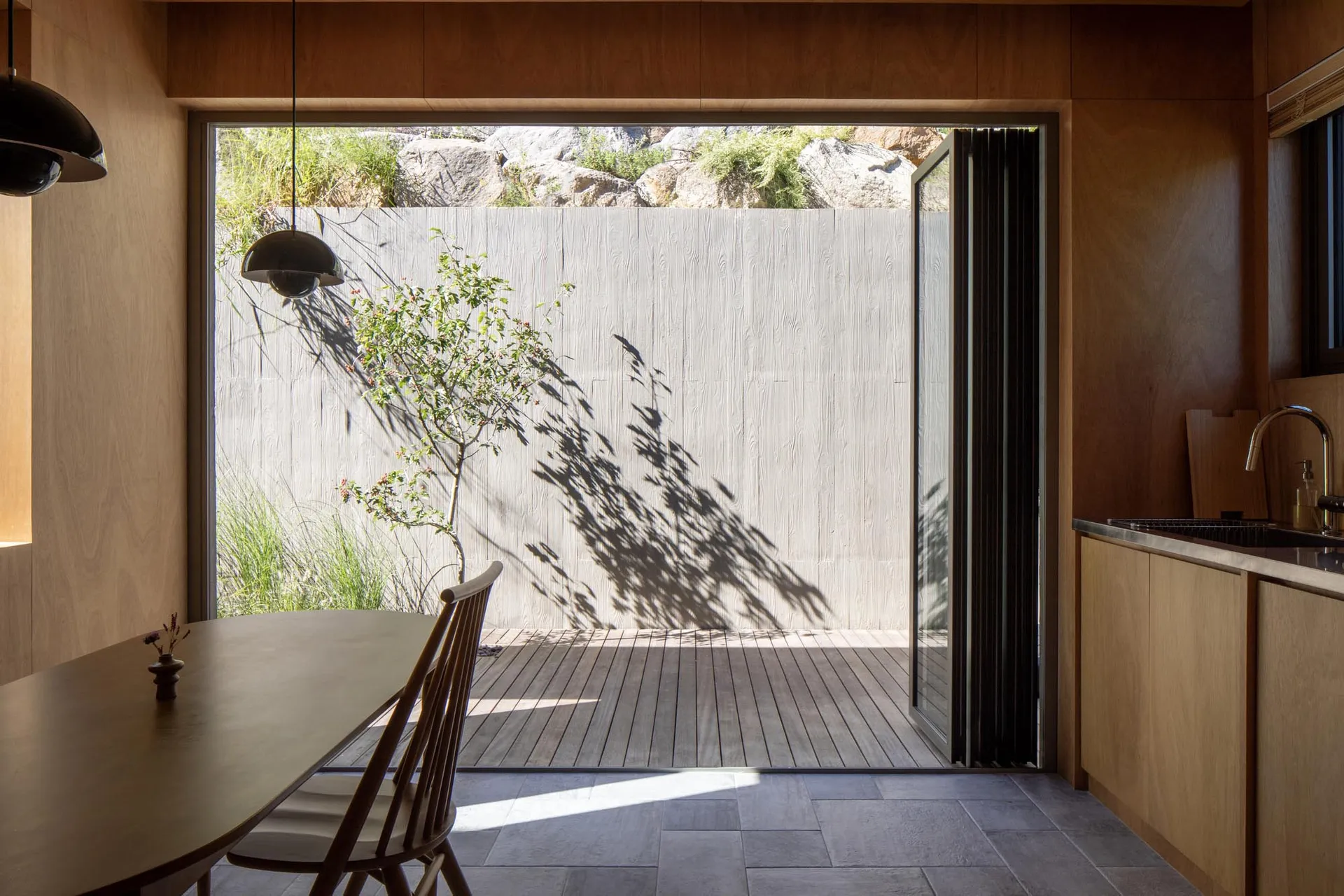 A minimalist residential kitchen featuring a wooden floor and a plant casting shadows on a wall.