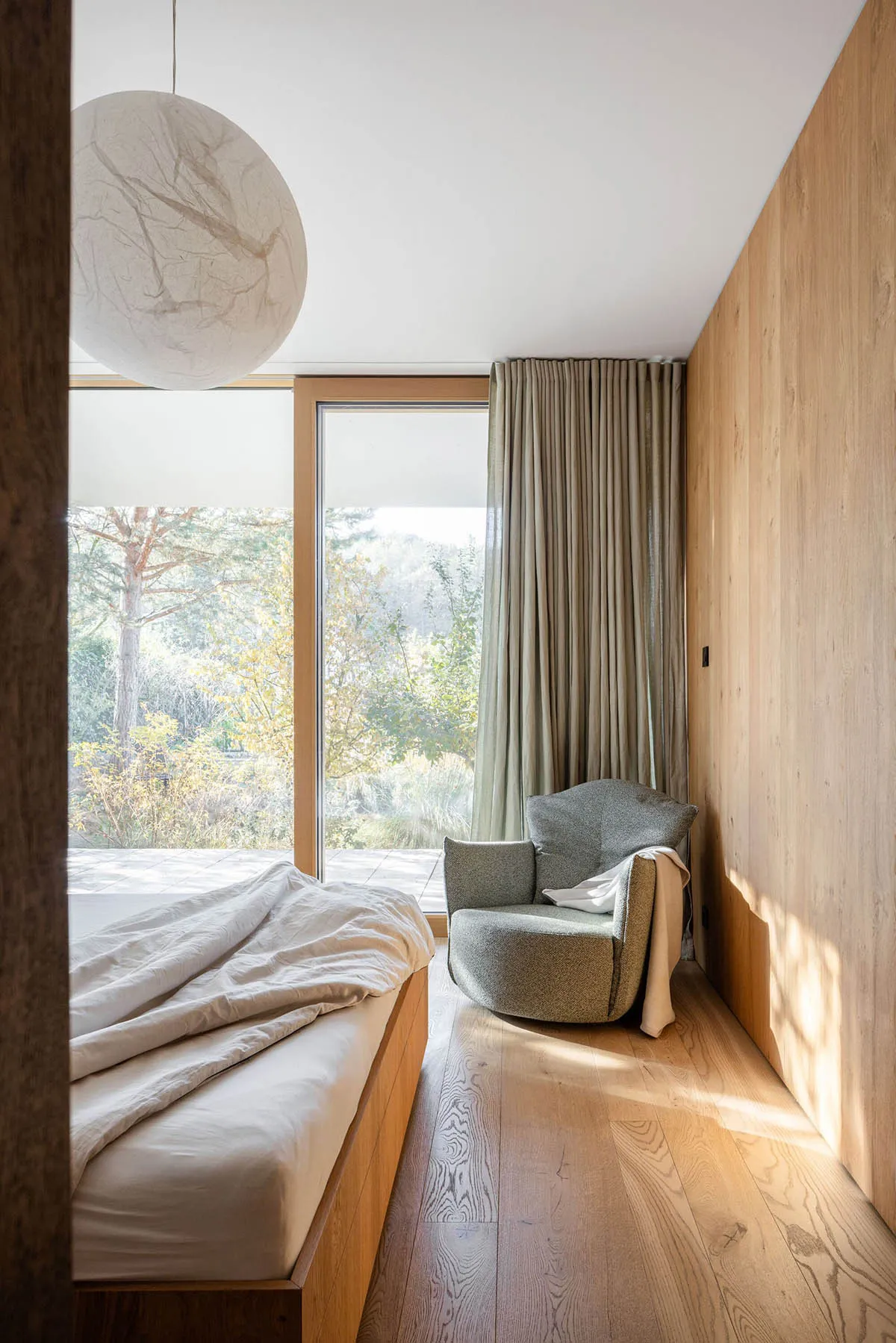 Modern minimalist bedroom with a wooden bed, a green armchair, and a large window overlooking nature.