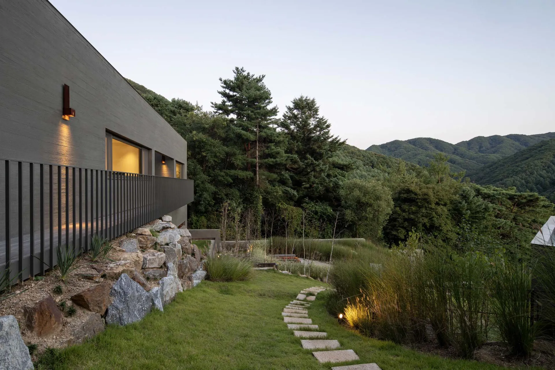 A modern residential exterior showcases a serene backyard garden pathway adorned with stones and surrounded by lush grass.