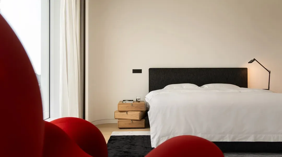 Modern minimalist bedroom featuring minimalist white fabric furnishings on a wooden bed platform, with a red curved chair and
