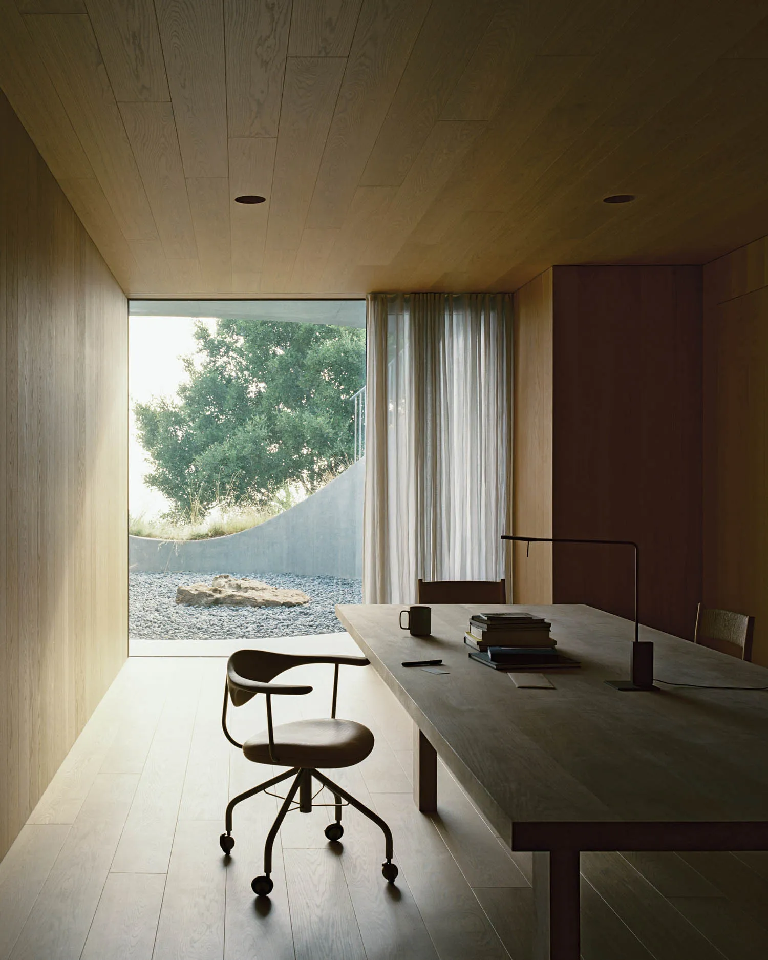 A minimalist office space featuring a wood table, chair, and bookshelf with books and a lamp, set against a serene outdoor vi