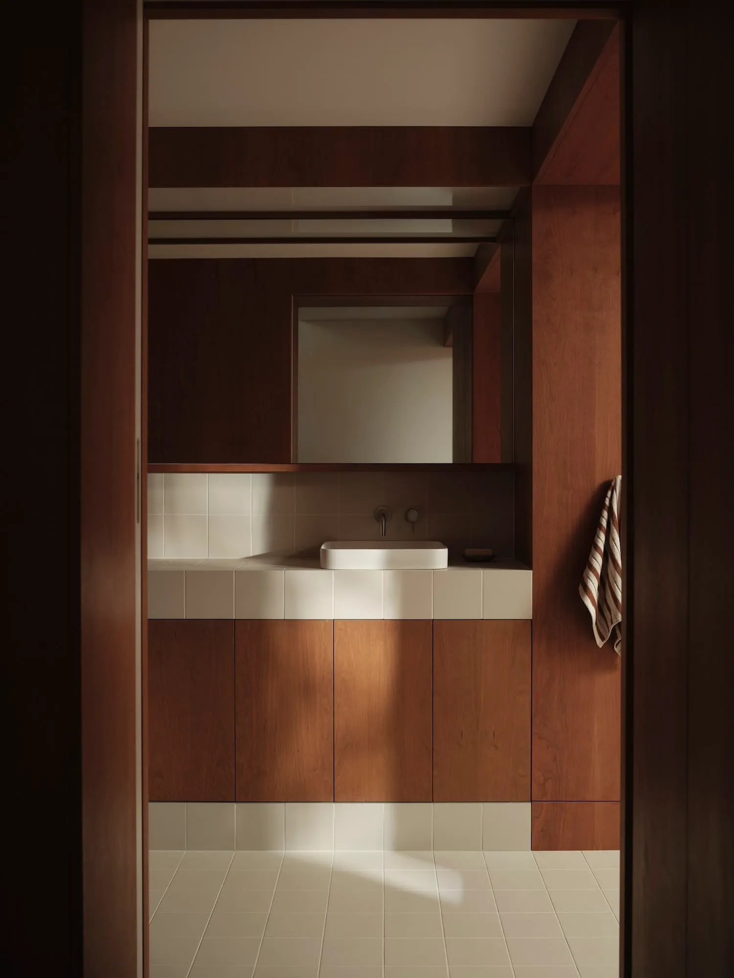 Modern bathroom featuring white tiled walls, a white countertop, a minimalistic sink, and wooden cabinets.