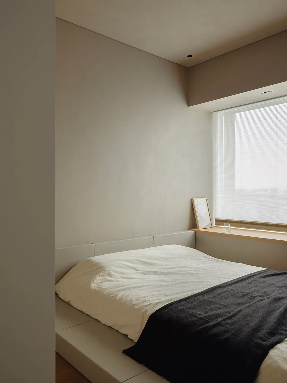 A minimalist bedroom with neutral-colored walls, bedding, and furnishings, featuring a window with closed blinds.