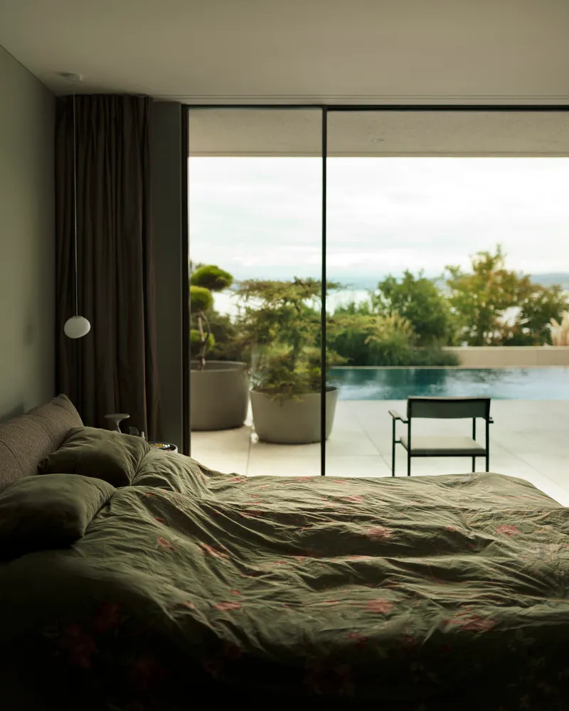 A serene minimalist bedroom featuring a bed with floral bedding, textured curtains, and a chair in front of a large window ov