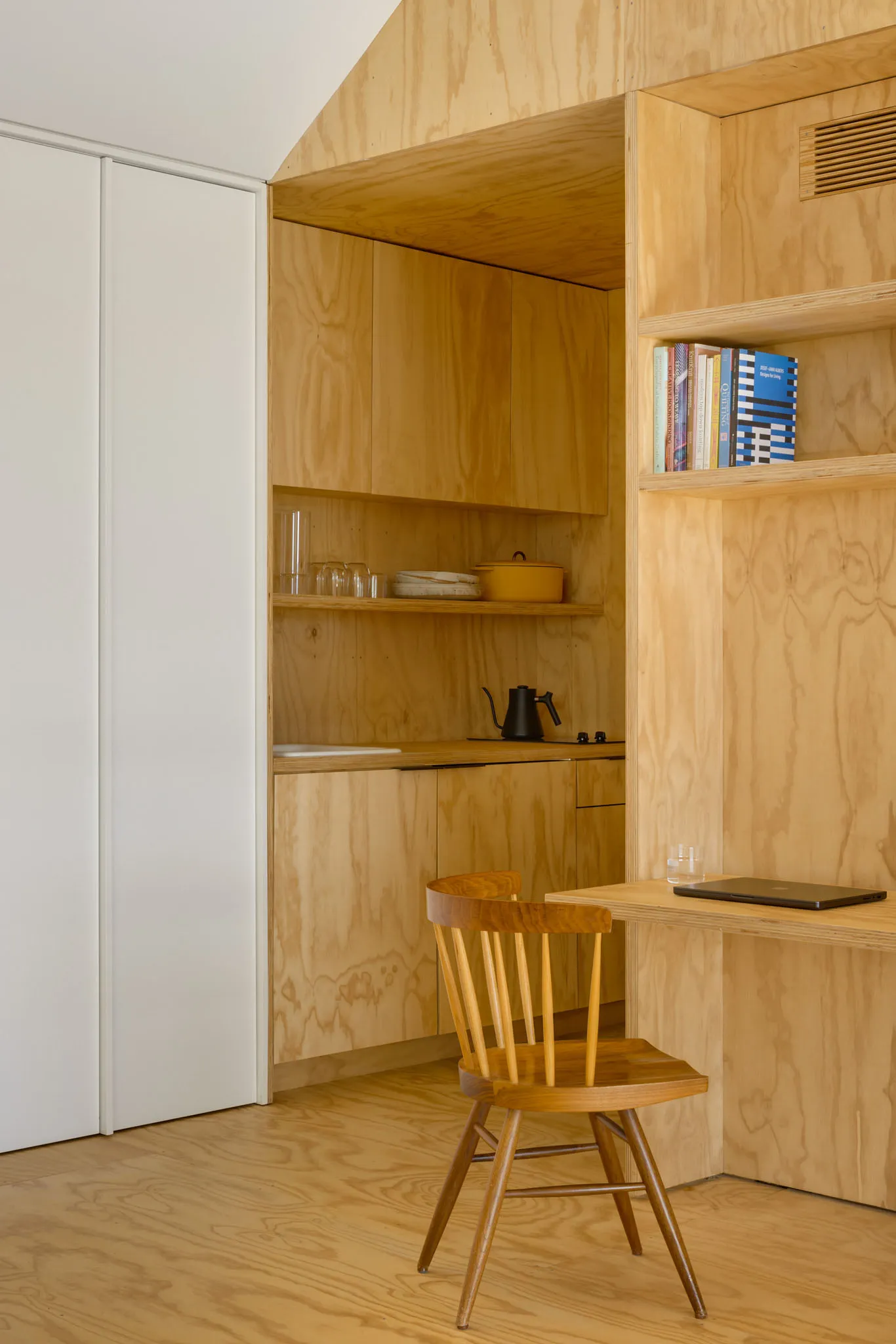 Minimalist kitchen with wood-paneled walls and built-in cabinets filled with glassware, cookware, and books.