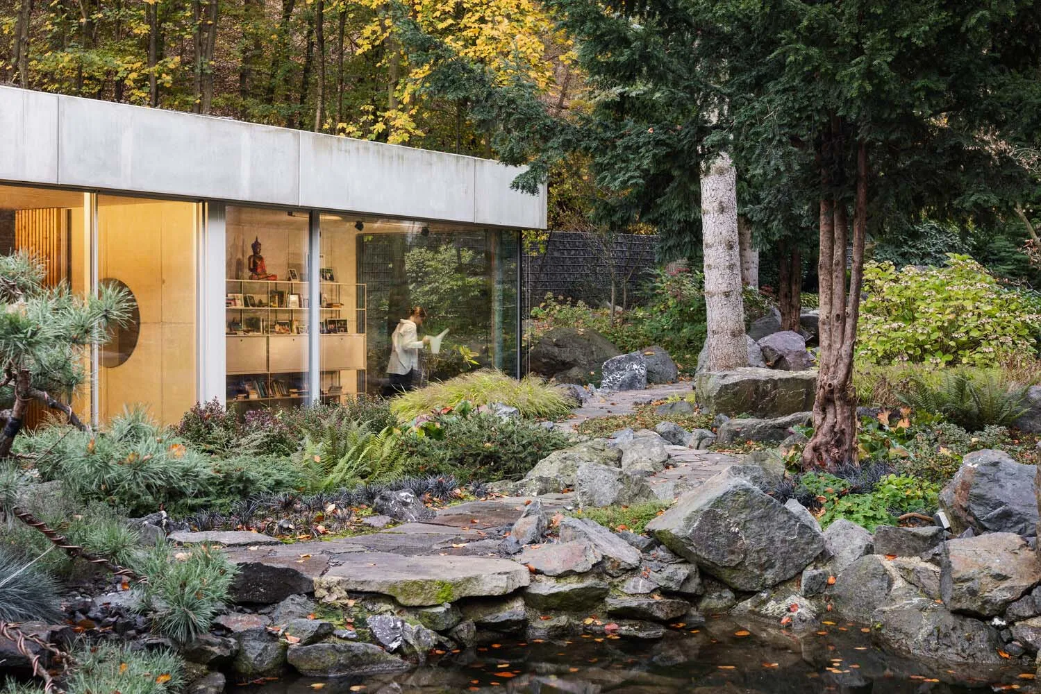 A minimalist garden featuring concrete structures, stone pathways, and glass elements surrounded by natural foliage and autum