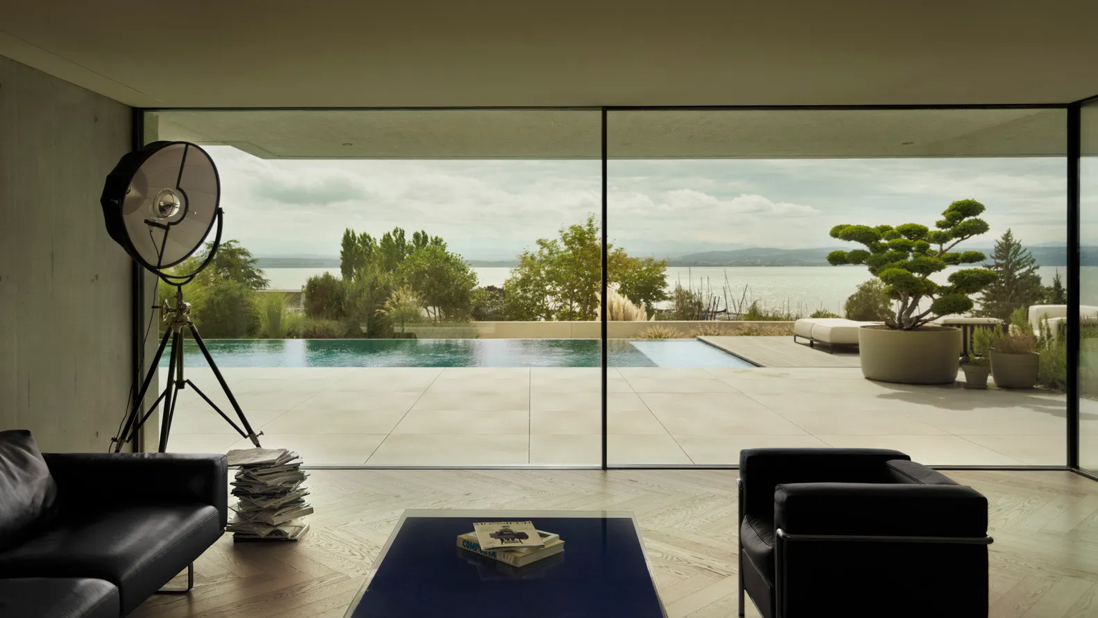 Modern minimalist living room featuring concrete walls and floor-to-ceiling glass windows overlooking a serene pool and natur