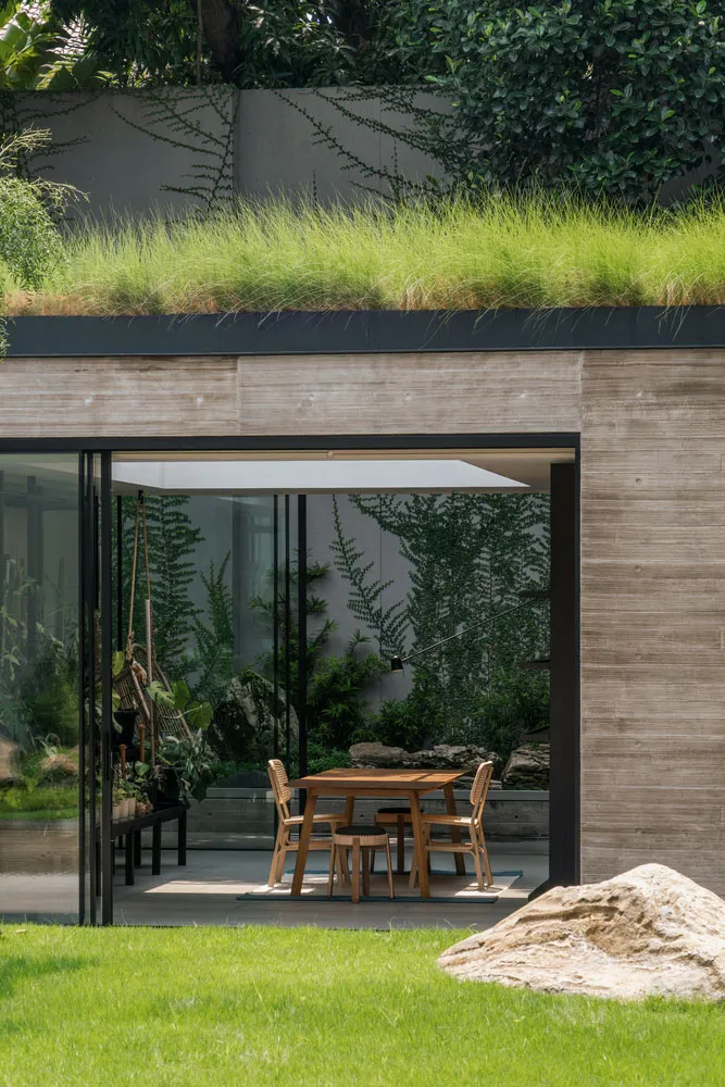 Modern garden extension featuring concrete walls, glass walls, rock formations, wooden furniture, and green plants.