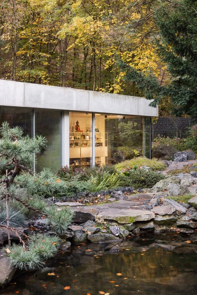 Modernist garden pavilion with concrete walls and glass windows surrounded by greenery and a pond.