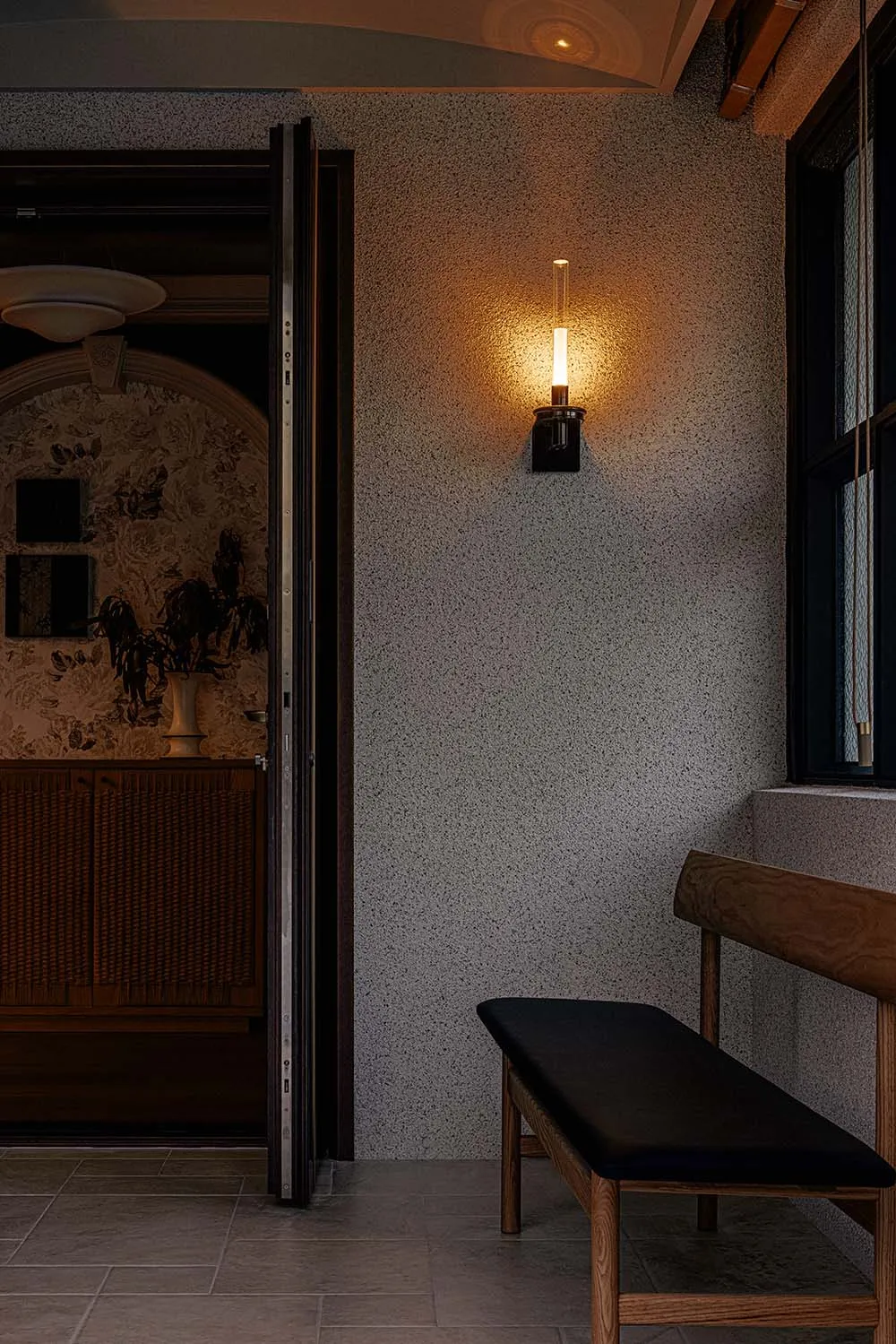 A modern residential hallway featuring a concrete textured wall, wooden bench, and warm lighting from a wall-mounted fixture.