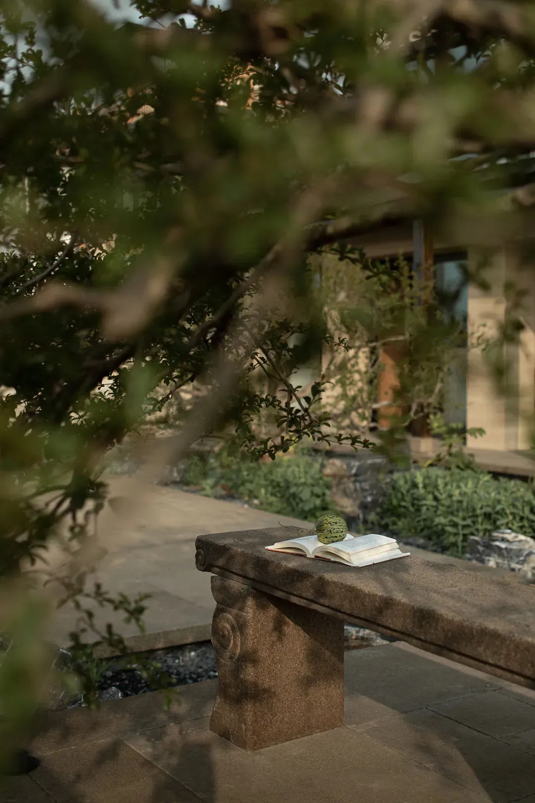 A stone bench with an open book and a fruit resting on top, set in an outdoor space with greenery and a house in the backgrou