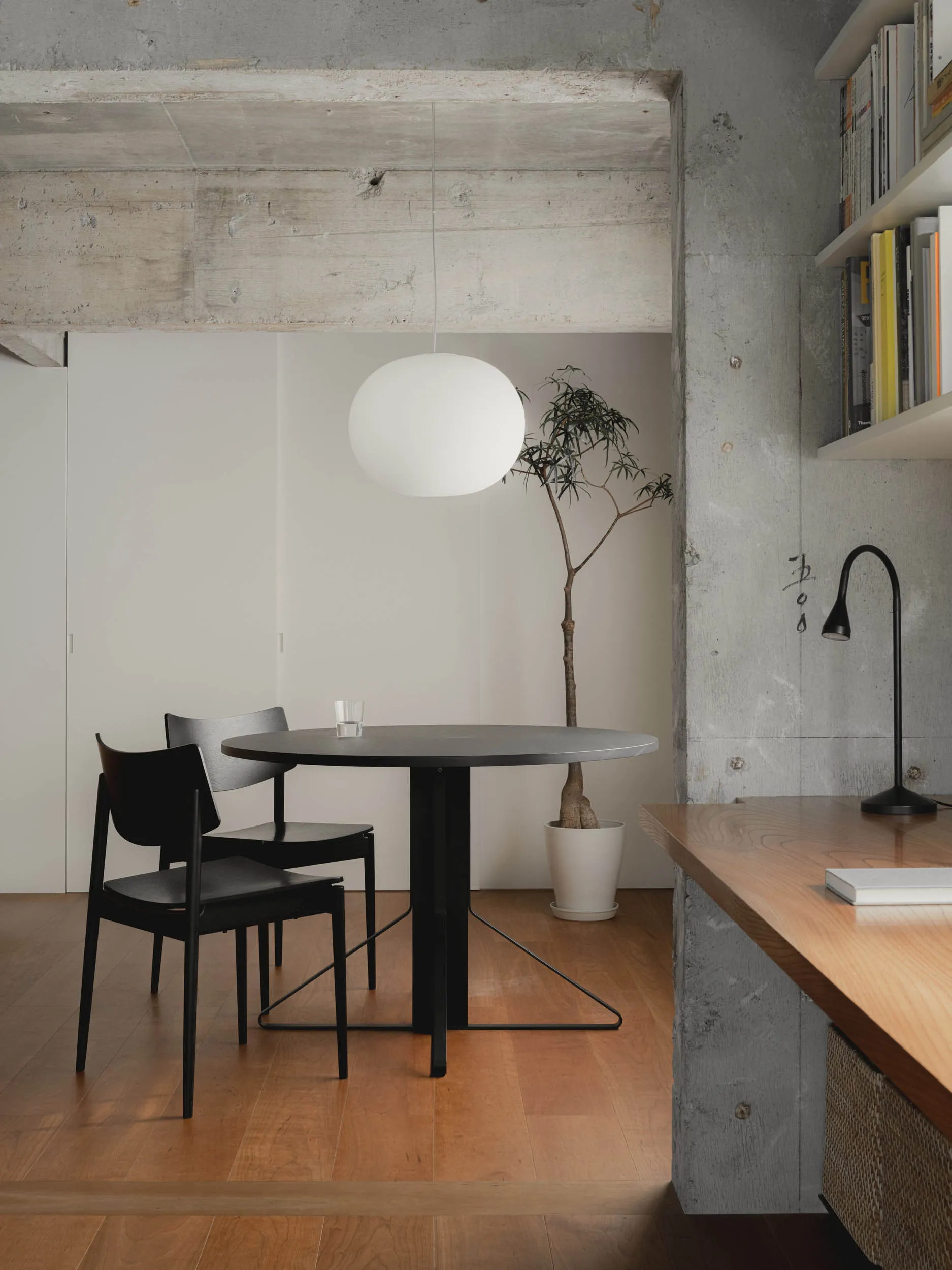 A minimalist dining area featuring a black table with white sphere pendant light, black chairs, and a tree in a white pot.