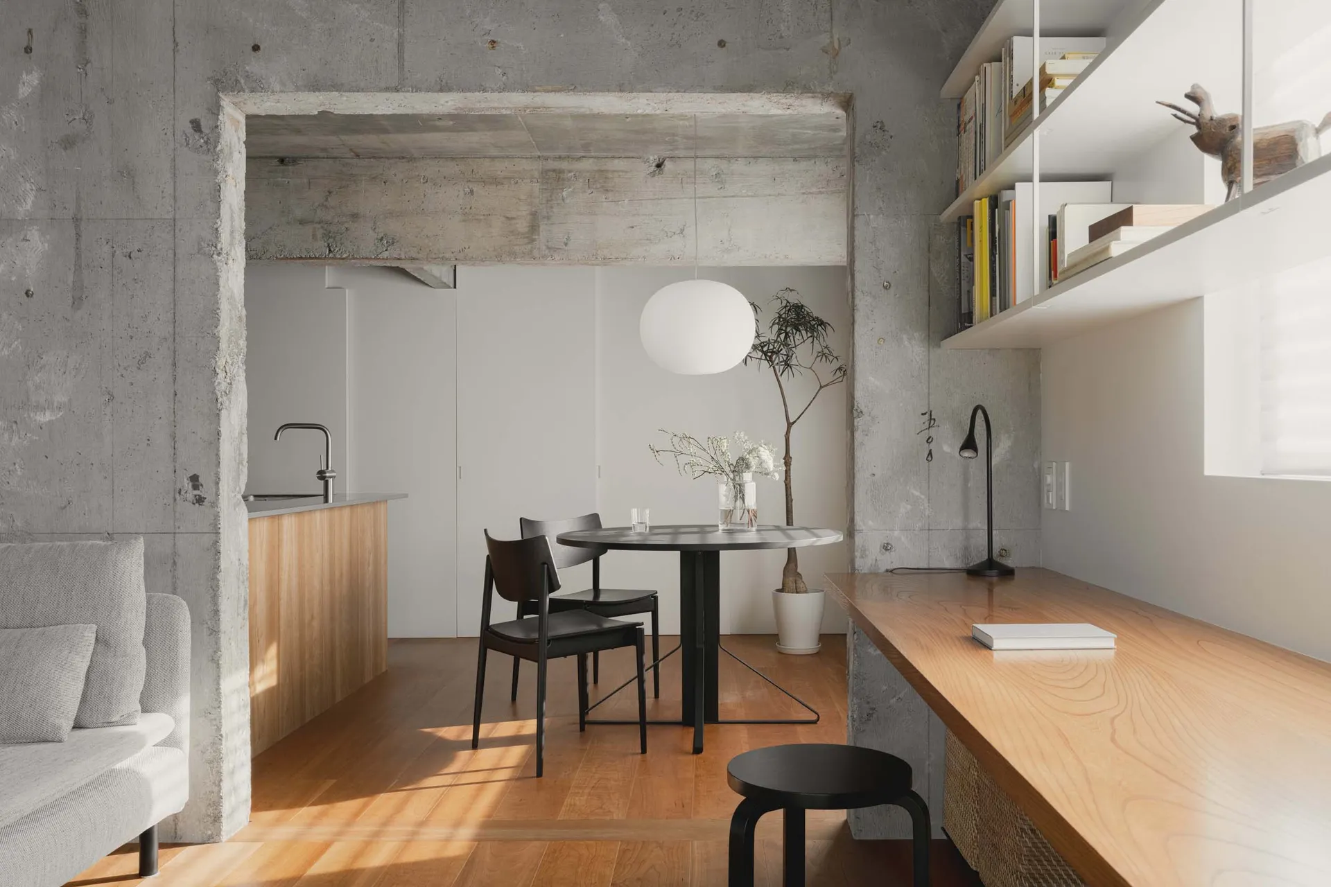 A minimalist living room with concrete and wood materials, featuring a round table, chairs, and a bookshelf.