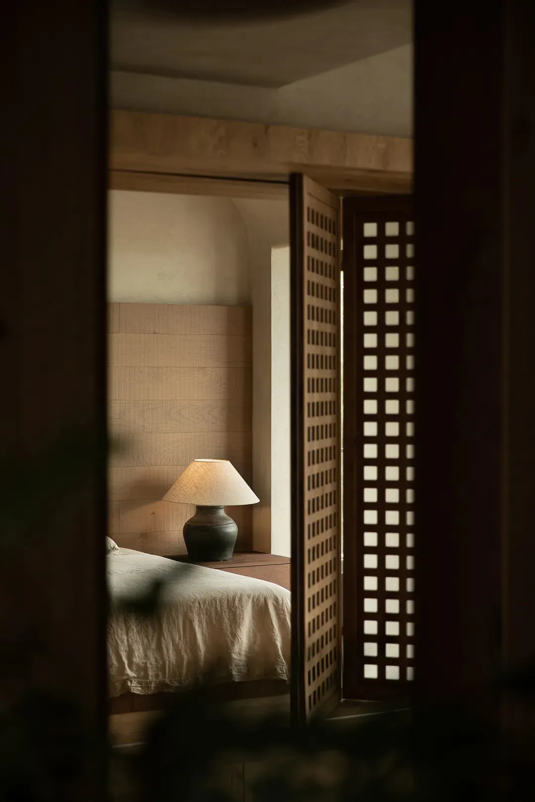 Minimalist bedroom featuring natural wood elements, with a cozy atmosphere created by a lit table lamp and a comfortable bed.