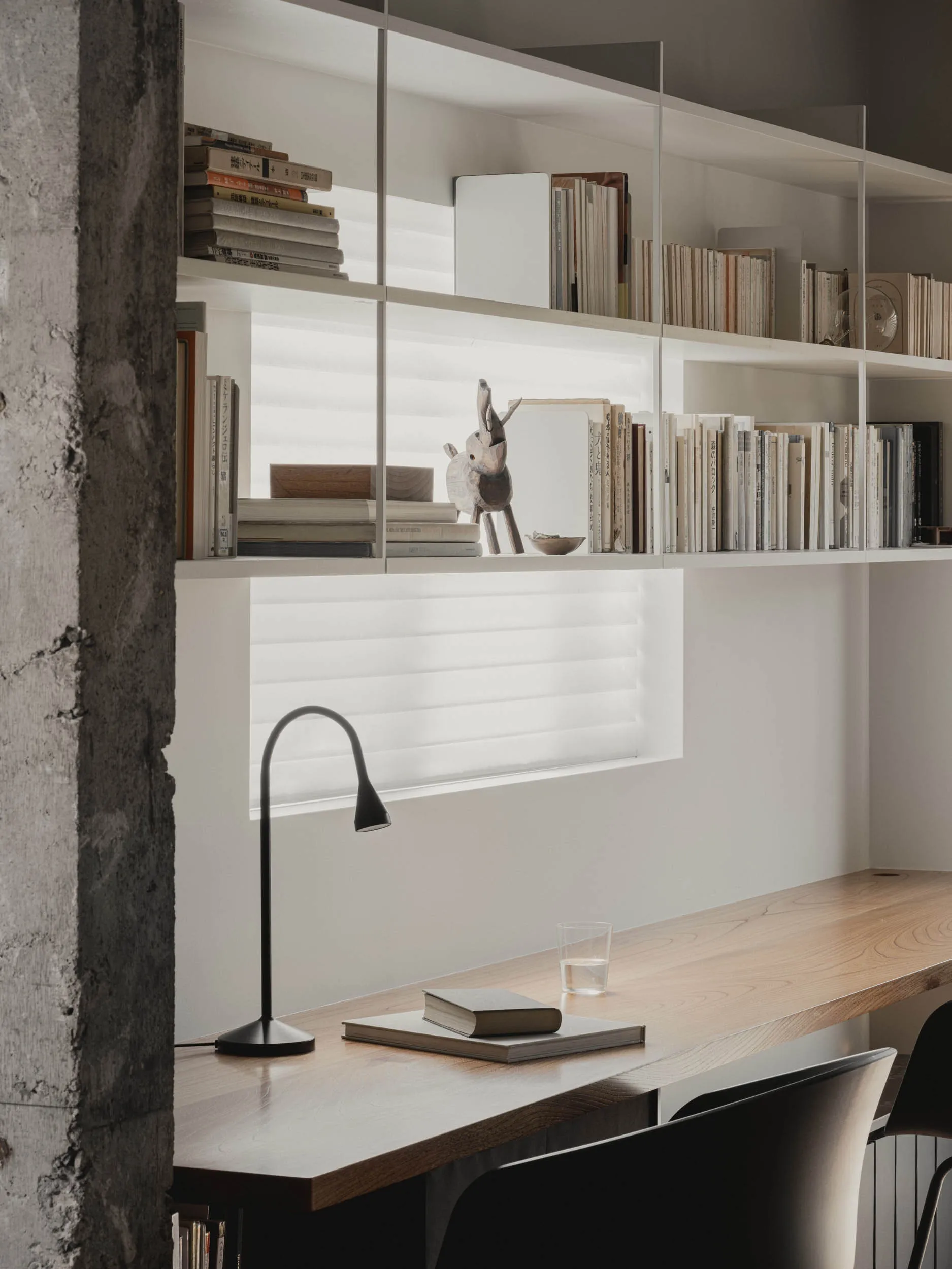 A minimalist office space with a wooden desk, bookshelf, and lamp, featuring a white background and books.