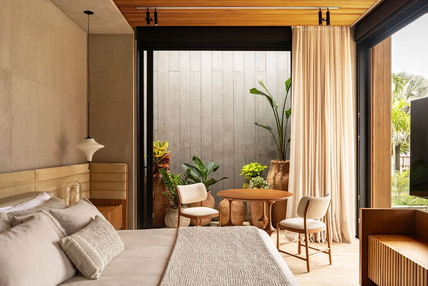 A minimalist bedroom with a bed, chairs, and a small table surrounded by potted plants, featuring hardwood floors and fabric