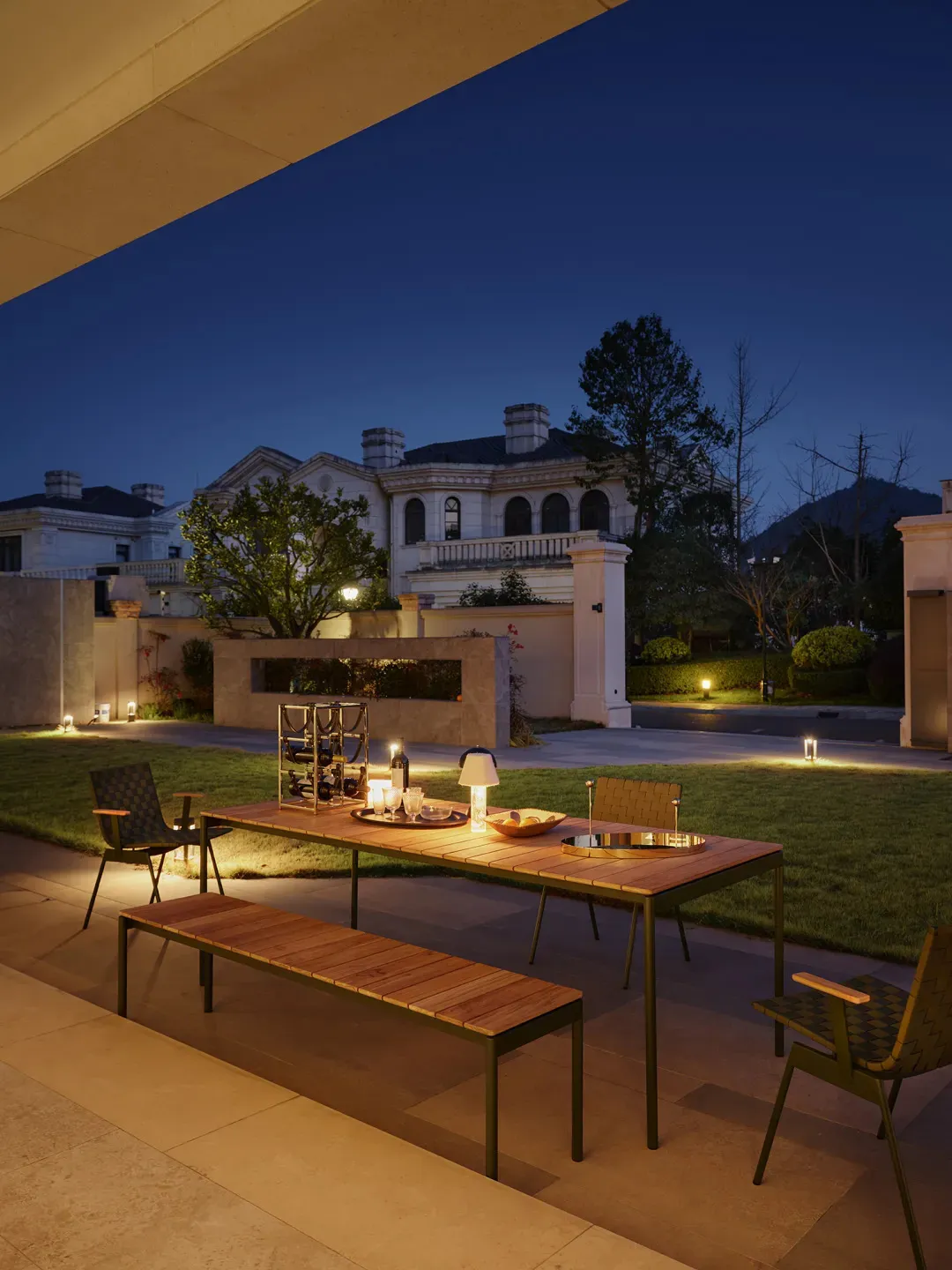 A residential exterior space with wooden and metal elements, featuring a table, chairs, and a bench under warm lighting.