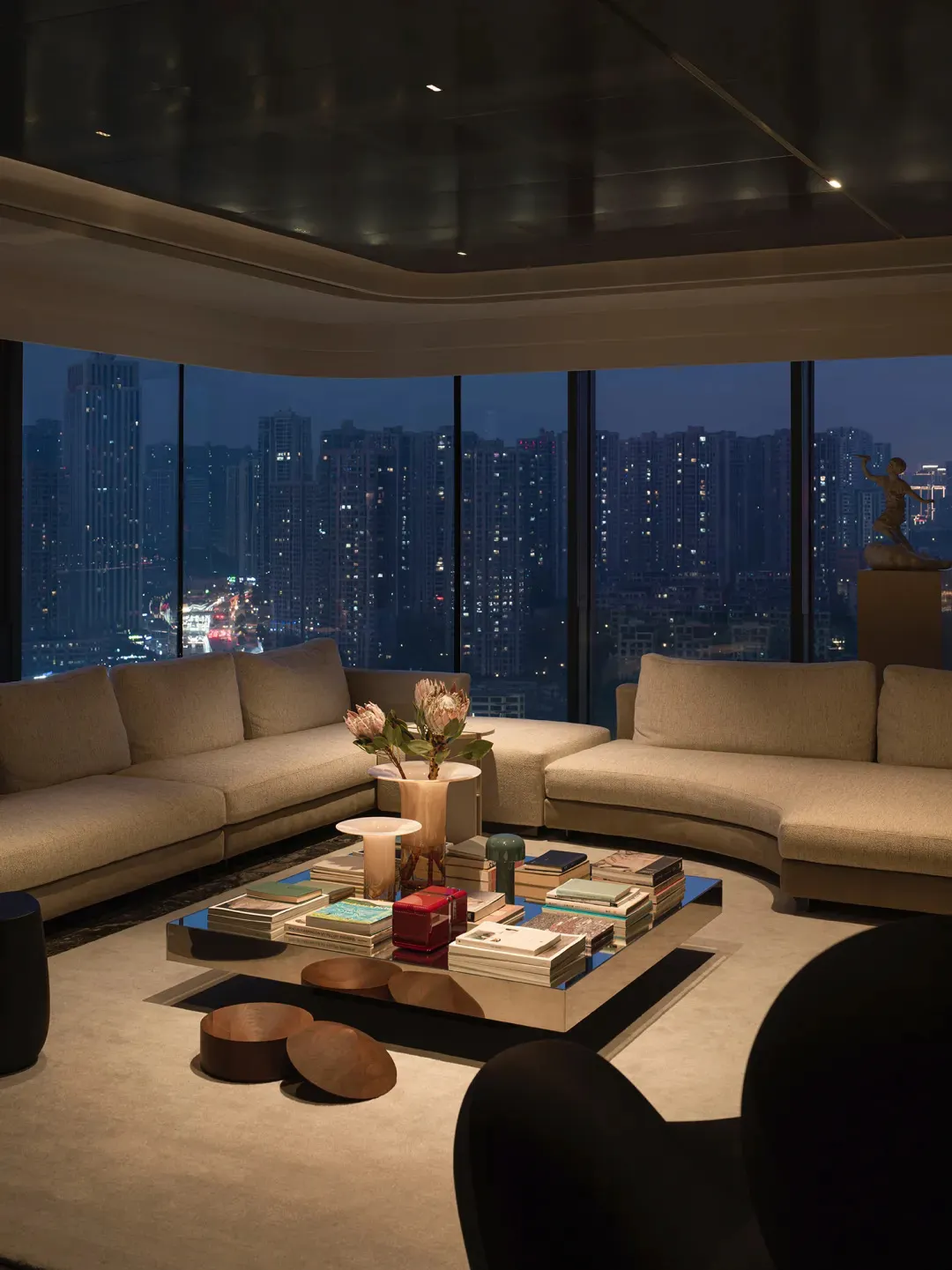 A modern minimalist living room with beige sofas, a square coffee table with books and decorative items, and a cityscape view