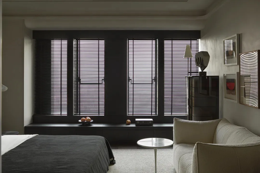Modern bedroom featuring metal blinds, a textile bed, a sofa, a lamp, and decorative pictures.