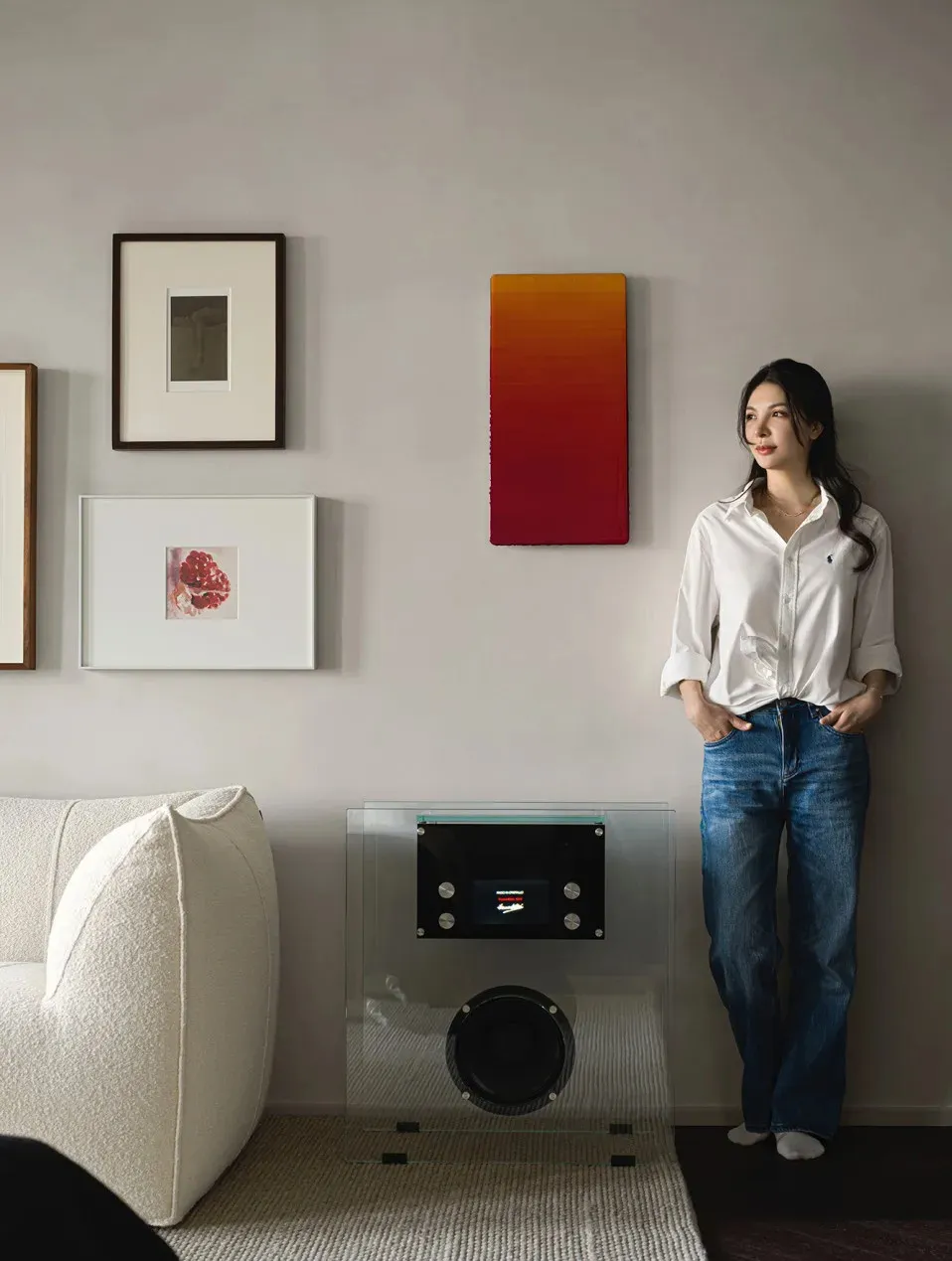 A living room with a modern glass side table, white couch, colorful artwork, and a woman standing against the wall.