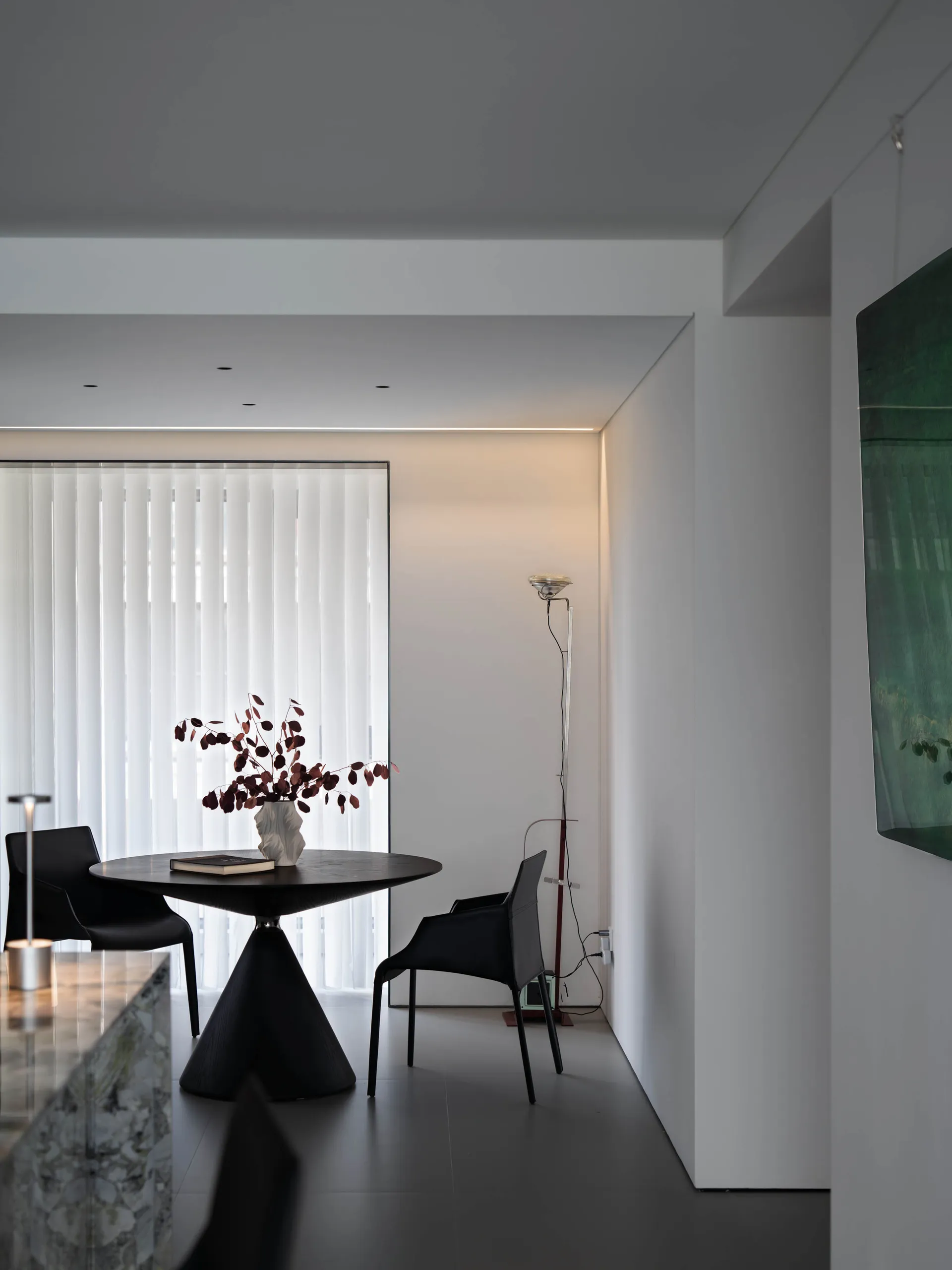 Modern minimalist dining area with a round glass table on a triangular concrete base and black chairs, featuring a vase with