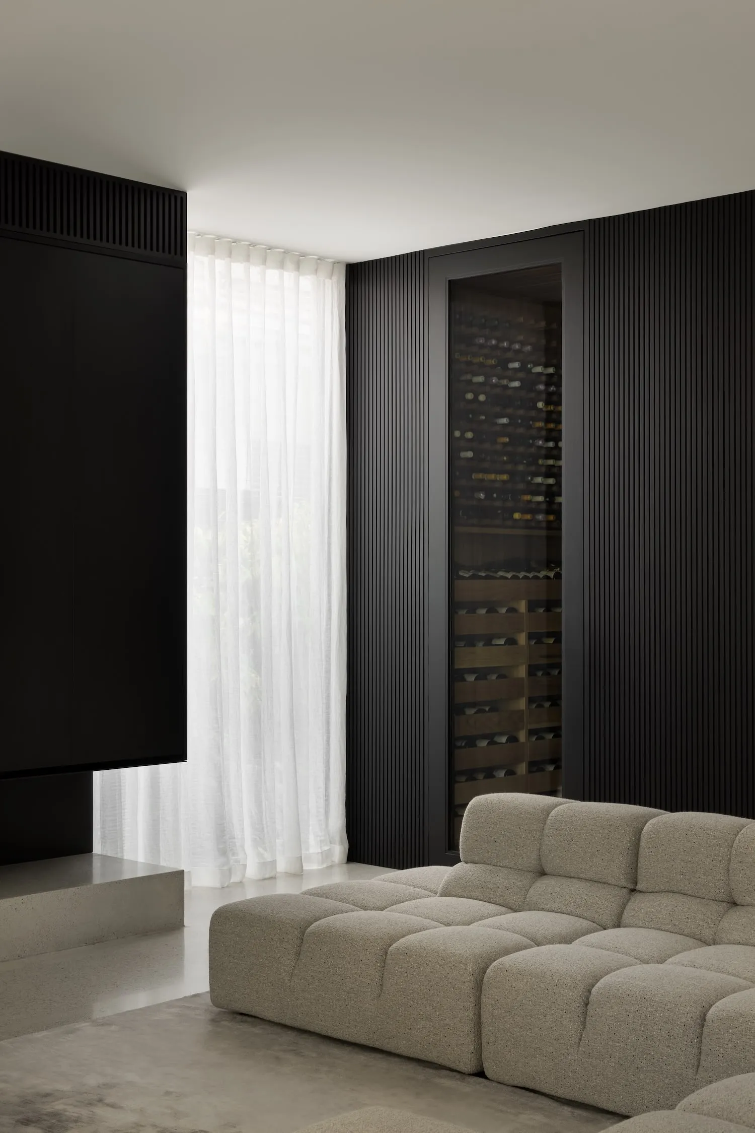 A modern living room featuring textured fabric sofas, a large screen TV, and a glass door wine cellar.