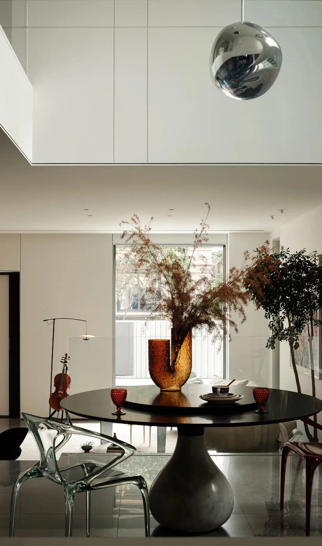 Modern dining room with white marble pedestal table, glass vase with dried plants, transparent acrylic chairs, and large glas