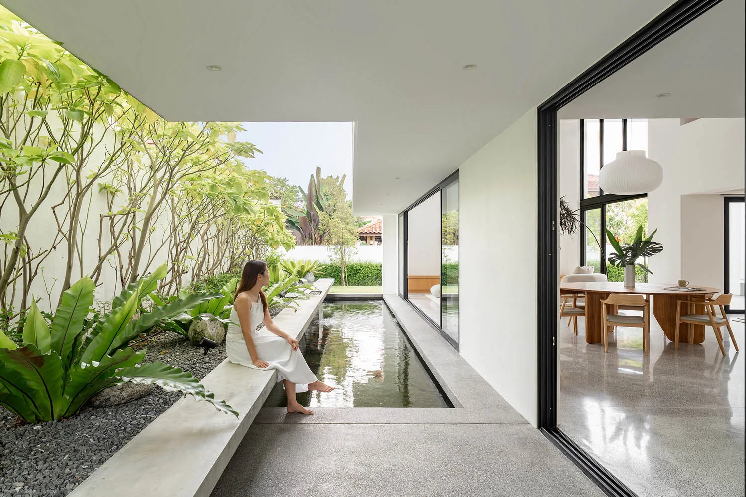 A woman in a white dress sitting in a minimalist indoor garden with a shallow pond and plants