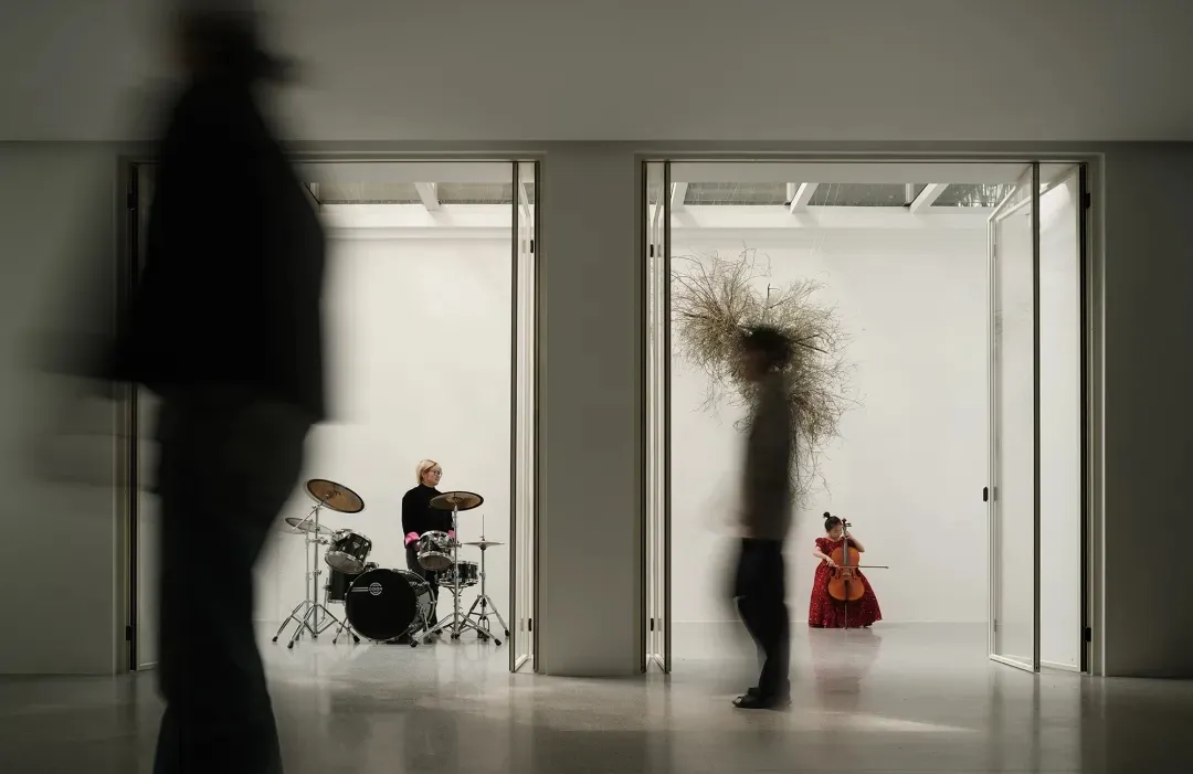 A minimalist interior photographic portrait depicting musicians in doors with a blurred figure walking by in the foreground.