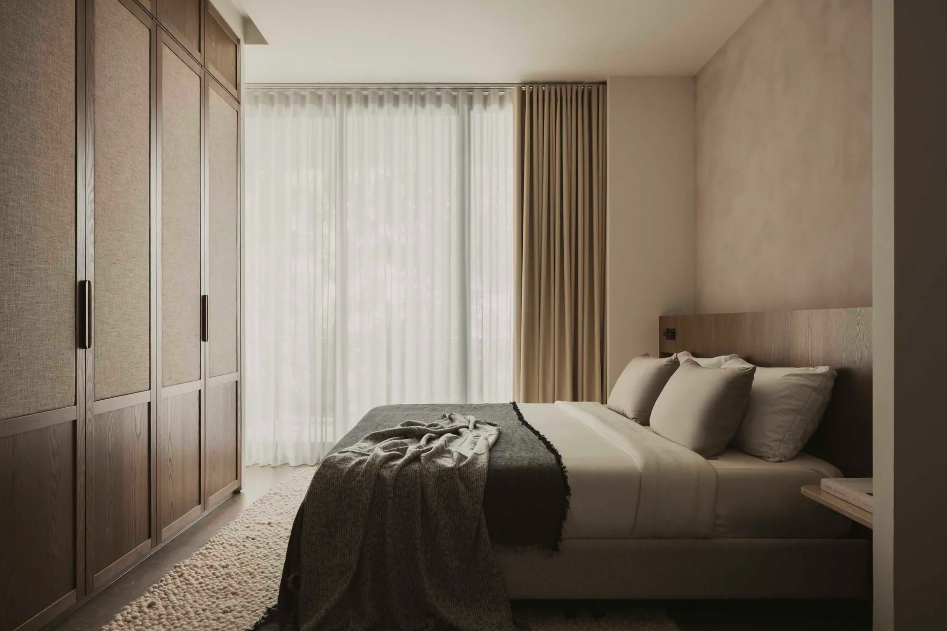 Minimalist bedroom featuring transparent window, wooden furniture, and beige fabrics.
