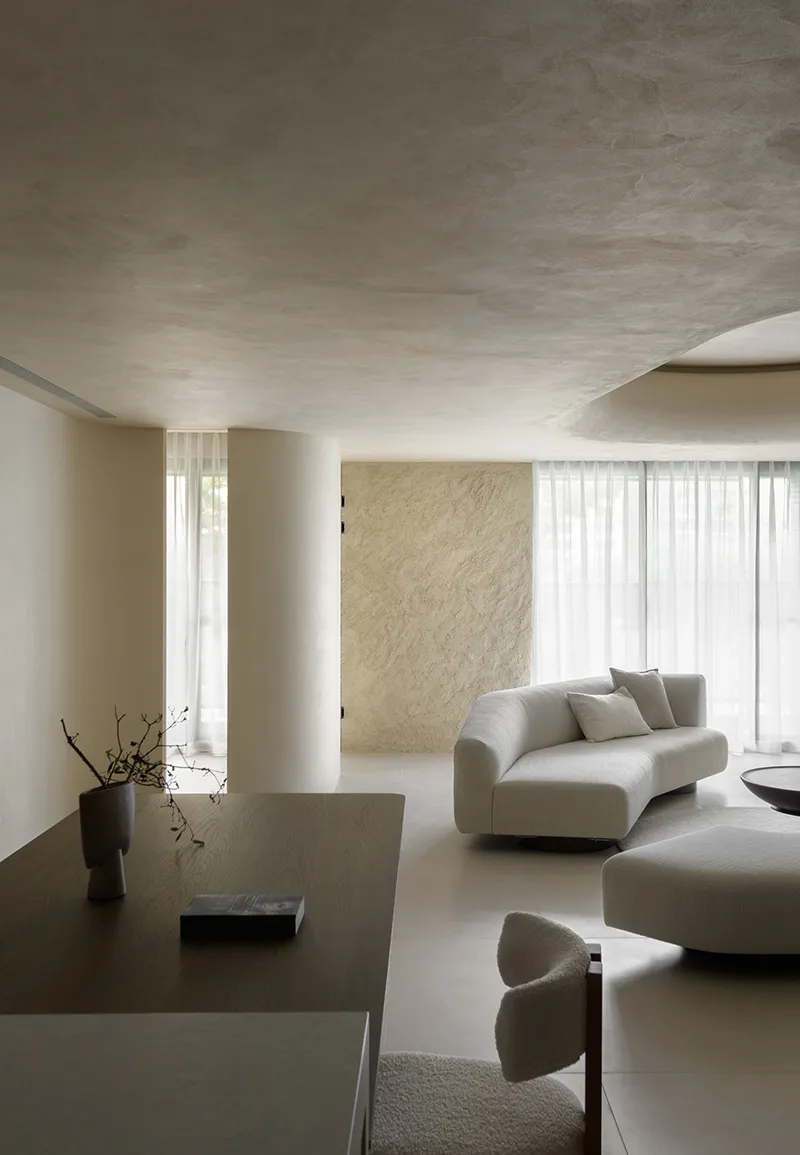 A modern minimalist living room with neutral-colored furniture and decor, featuring a round sofa, a wooden dining table, and