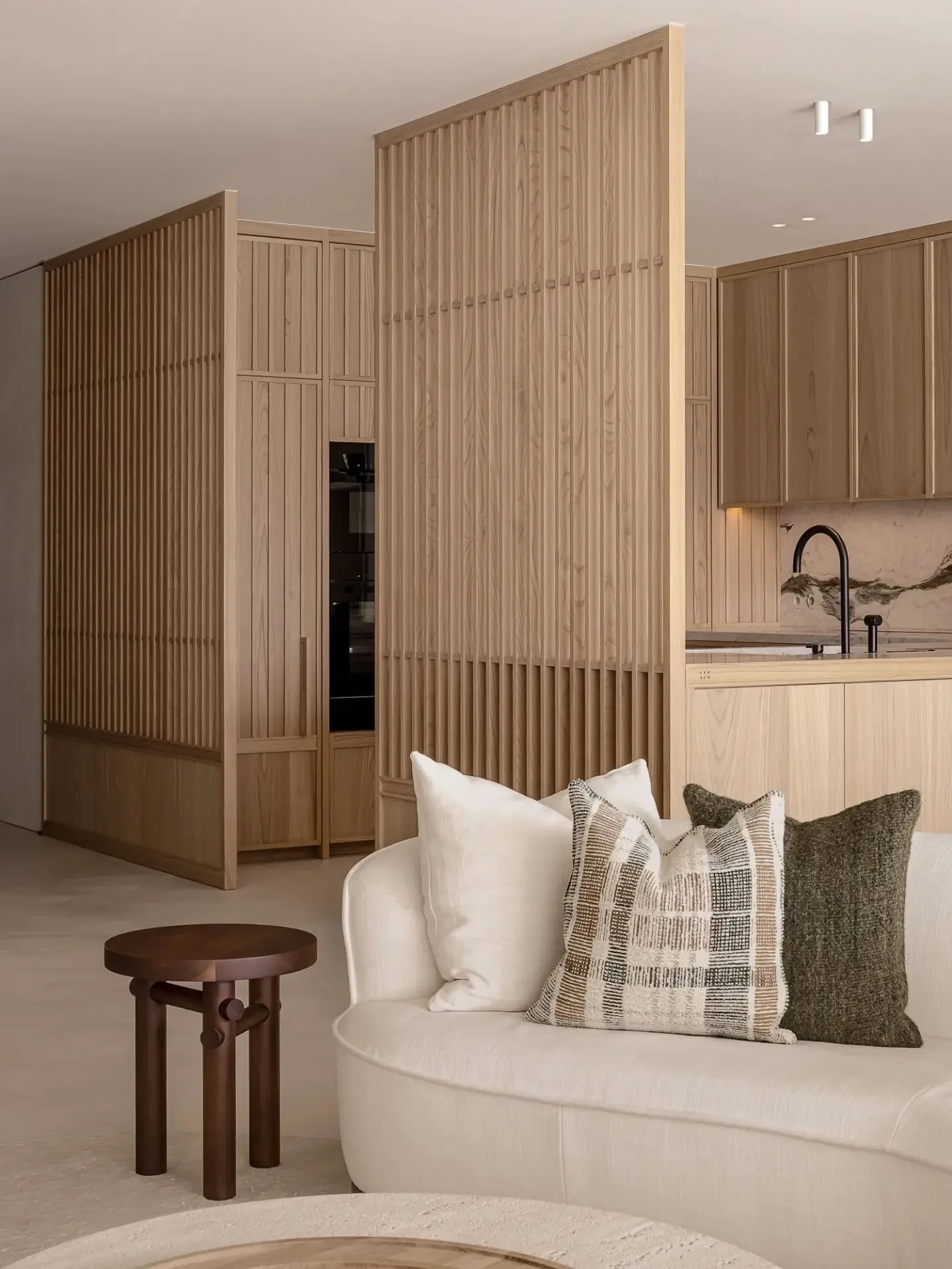 A modern residential interior featuring a white sofa with patterned and solid pillows, wooden furniture, and a kitchen area w