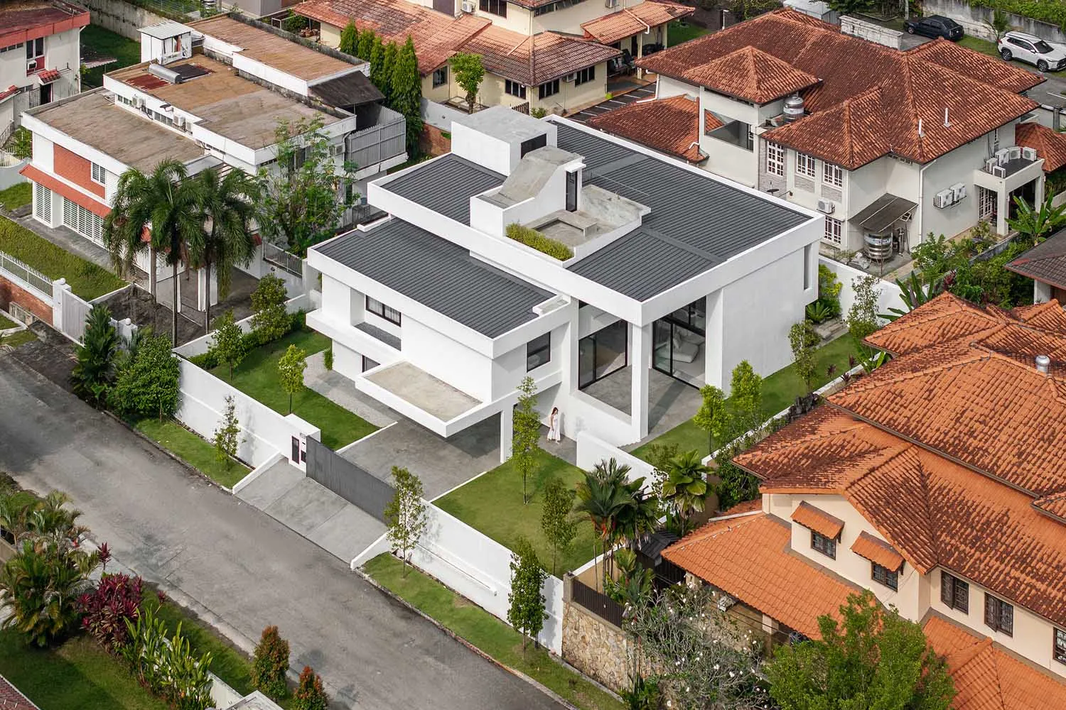A modern white house with a white roof surrounded by other houses with brown tiled roofs and palm trees.